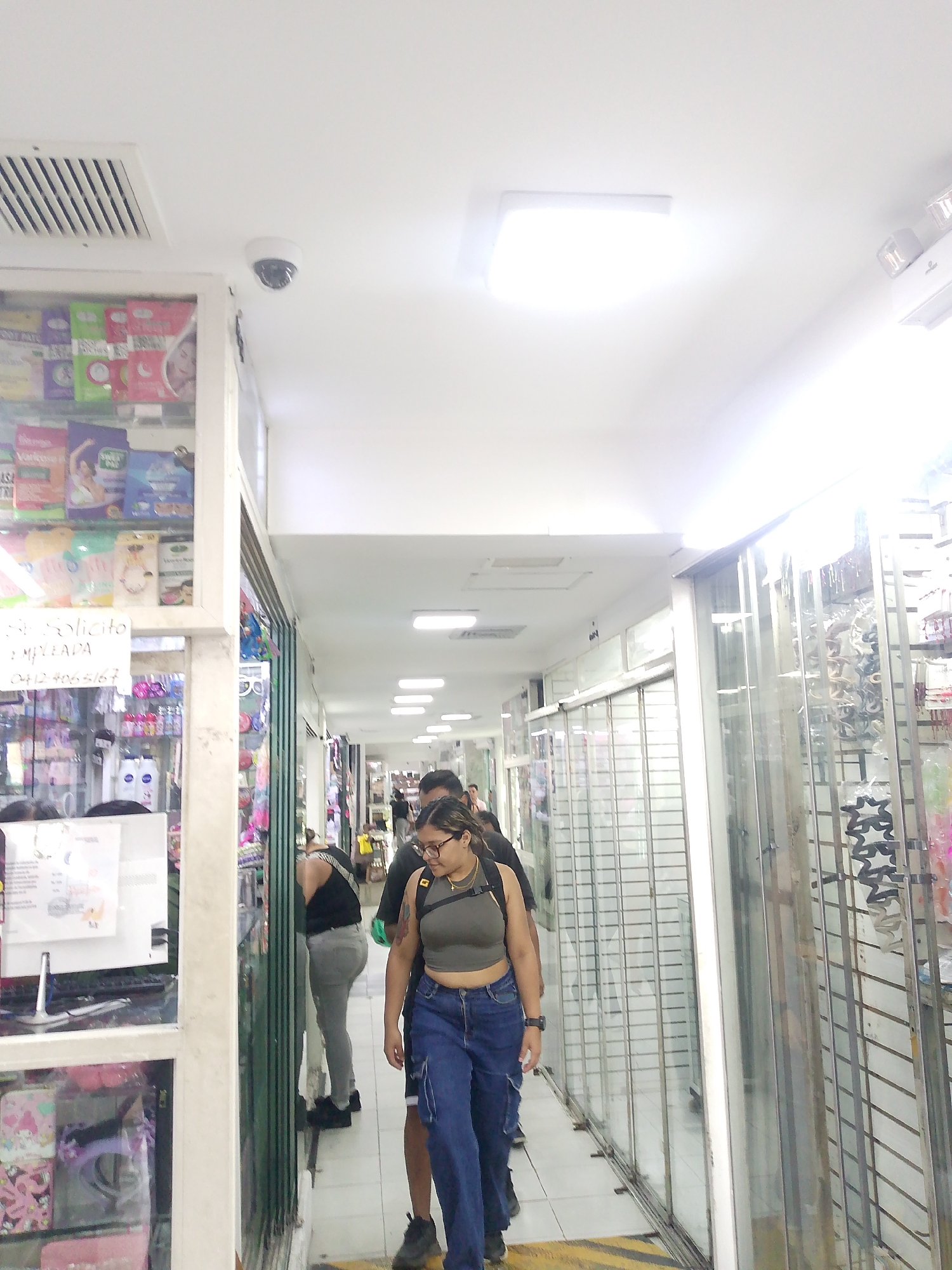 The image shows an indoor commercial space, likely a shopping arcade or market, in Caracas, Venezuela. The architecture consists of a long, brightly lit corridor with shops lining either side. The shops are enclosed with metal security grills and glass displays showcasing various merchandise, including cosmetics and accessories. Three people are walking along the corridor; one wearing a grey top, jeans, and glasses is in the foreground, with two others behind them. A security camera is mounted on the ceiling. The floors are tiled, and there are yellow and black hazard stripes near the shop fronts. A sign "Se Solicito Empleada 0412 406 5167" is visible on a shop display.