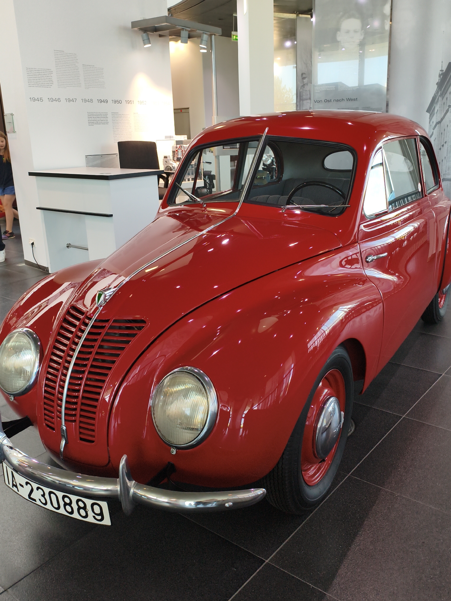 The image depicts a red vintage car on display in a museum. The car is the primary subject, positioned in the center of the frame, with its front facing slightly towards the right. It is a two-door sedan with a rounded body, prominent headlights, and a grille with horizontal bars. A license plate with the inscription "IA-230889" is attached to the front bumper.
In the background, a white wall is visible, with a timeline of years marked from 1945 to 1952. Next to the wall is a white desk with a black chair behind it. Further in the background, large posters with black and white images are displayed, one featuring a portrait and text that reads "Von Ost nach West." The flooring is dark, tiled, and appears to be polished.
To the left of the frame, a young woman with blonde hair is partially visible, wearing a dark top and blue shorts. She is walking away from the camera.
This scene is located in the Audi Museum Mobile in Ingolstadt, Germany. The museum showcases the history of Audi and its predecessors.