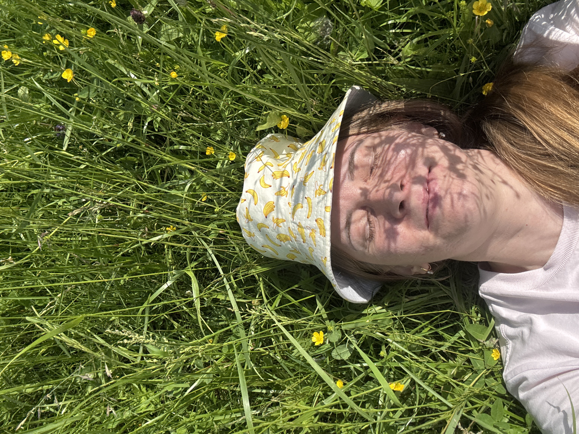 The image depicts a young Caucasian woman lying in tall green grass, with her eyes closed and a relaxed expression on her face. She is wearing a white t-shirt and a bucket hat adorned with a banana pattern. The sun casts dappled shadows on her face, suggesting it's a bright, sunny day. The surrounding environment is a meadow or field, with numerous small yellow flowers interspersed among the grass. The overall scene conveys a sense of peace and enjoyment of nature. The location is identified as the city of Курган, Russia.