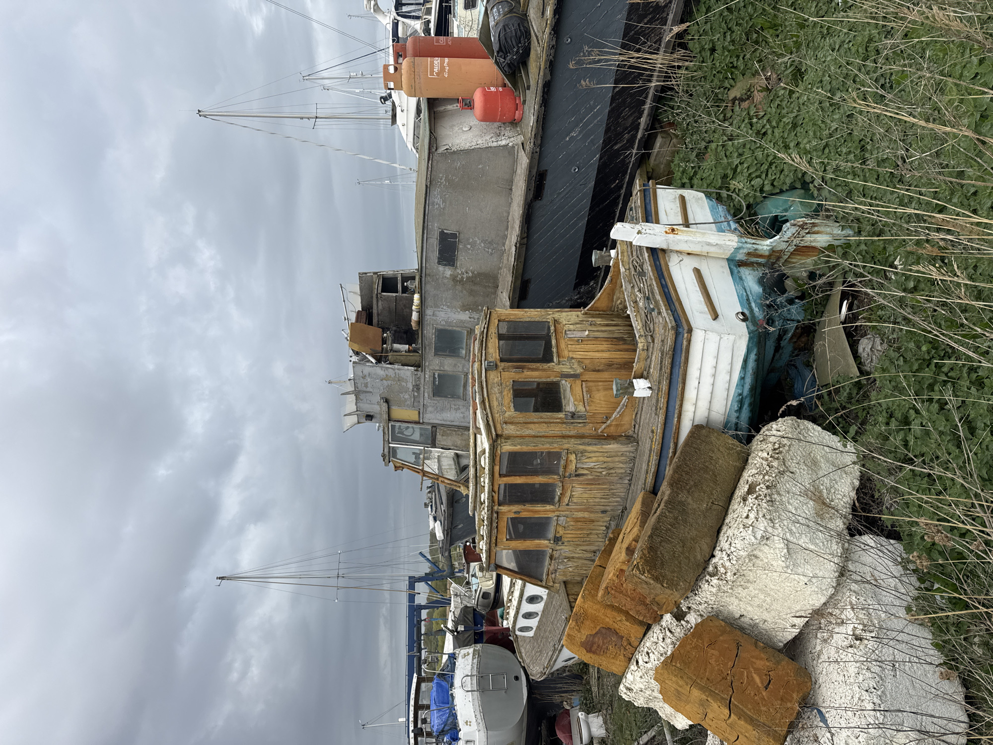 This image depicts a cluttered boatyard or coastal storage area under an overcast sky. In the foreground, a heavily weathered wooden boat with peeling paint and numerous broken window panes rests on land. Adjacent to it, a pile of large, irregular blocks, appearing to be a mix of concrete and aged timber, is visible. The ground around these objects is covered in dense green vegetation and tall, dry grasses.
Behind the weathered wooden boat, a larger, dark-hulled vessel is prominent, with two orange propane tanks on its deck. Visible text on one of these tanks reads "CALORGAS" and "47 kg Propane". A smaller red propane tank is also present on the deck. Further into the background, numerous sailing yachts with tall, slender masts are visible against the cloudy sky, some appearing to be on land, others possibly in the water. Many of the boats show signs of age, disrepair, or long-term storage.
The weather is distinctly overcast, with a uniform grey, cloudy sky dominating the upper portion of the image. The time of day appears to be daylight, given the overall illumination, but without direct sunlight. There is no visible human activity. The scene evokes a sense of disuse and the passage of time, characteristic of a working or disused boatyard, consistent with the provided location context of East Suffolk, United Kingdom.