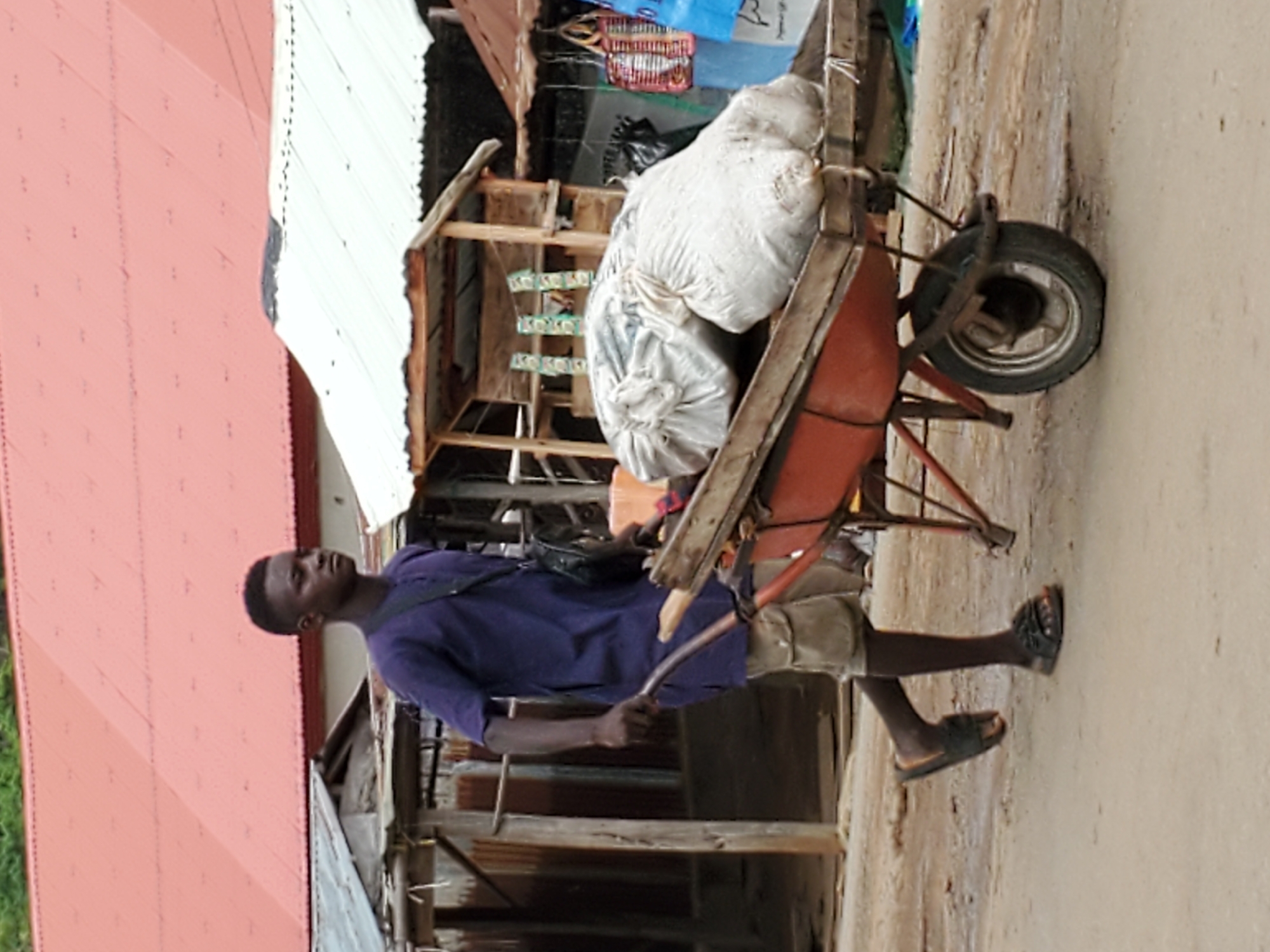 A man walks on a dirt road in Mararaba, Nigeria, pushing a red wheelbarrow loaded with two large white sacks. He wears a purple short-sleeved shirt, khaki shorts, and black sandals, and a black bag is slung across his shoulder. The wheelbarrow has a single front tire and wooden handles. Behind him are corrugated metal buildings and a structure with hanging green decorations. To the left is a building with a red roof. The ground is dirt.