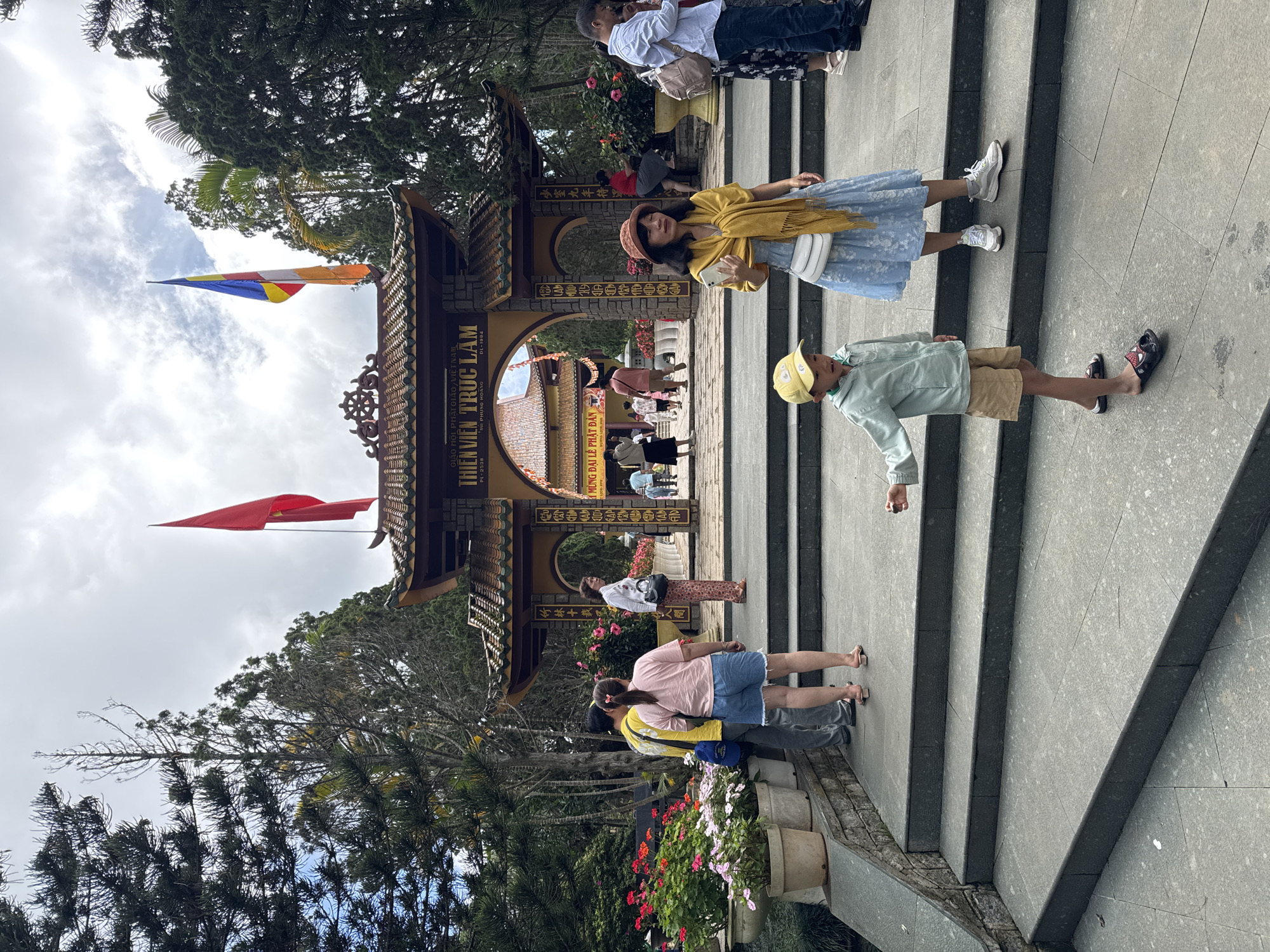 This image captures the entrance gate of Thiền Viện Trúc Lâm (Truc Lam Zen Monastery) in Da Lat, Vietnam, under an overcast sky. The prominent feature is a traditional Vietnamese archway constructed from stone and dark wood with a tiled roof, positioned centrally in the midground. Inscriptions in Vietnamese are visible on the archway, including "GIÁO HỘI PHẬT GIÁO VIỆT NAM" (Buddhist Sangha of Vietnam) and "THIỆN VIỆN TRÚC LÂM" (Truc Lam Zen Monastery). A red Vietnamese flag with a yellow star is mounted on a pole to the right of the gate, while a multicolored Buddhist flag (blue, yellow, red, white, orange stripes) is on a pole to the left. The gate is flanked by dense green foliage, including tall, dark evergreen trees and other tropical plants. The ground in the foreground consists of wide, light-grey granite steps leading up to the gate, with a flat paved area at the lowest level. A stone-faced planter wall along the left side of the steps holds multiple potted plants with colorful flowers, predominantly red, pink, and white. Numerous individuals are present on and around the steps. In the immediate foreground, a young boy stands on the lowest step, facing slightly left, wearing a yellow baseball cap, a light mint-green hooded jacket, tan shorts, and black slide sandals with a red Spider-Man motif. To his right and slightly higher on the steps, a woman stands facing towards the right side of the frame. She wears a wide-brimmed reddish-brown hat, a mustard-yellow shawl draped over a light blue dress with a white floral pattern, white sneakers, and a white crossbody bag. She holds a white smartphone in her left hand. To the left, in the midground, a man with his back to the camera wears a yellow t-shirt, grey pants, and a blue shoulder bag. He stands next to a woman in a light pink t-shirt, blue denim shorts, and pink slide sandals, also with her back to the camera. To the right of the gate, another woman stands facing right, dressed in a white long-sleeved shirt, patterned dark pants, and black shoes, carrying a black handbag. Further to her right, a man in a light blue collared shirt and dark pants, with a light brown backpack, looks down at a phone. A woman in a floral dress is partially visible beside him. Inside and beyond the archway, several other people are visible,