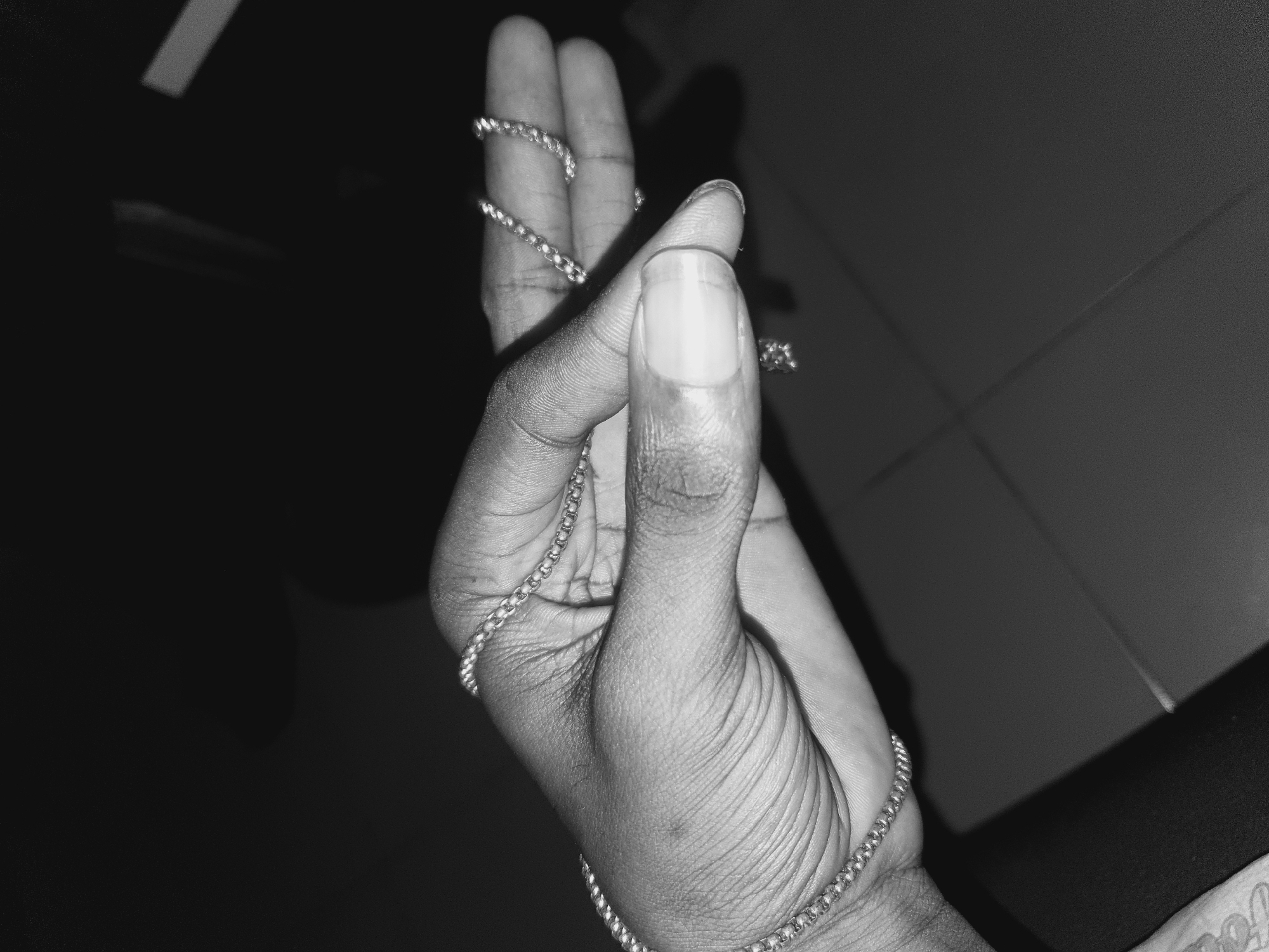 A black and white, close-up photograph shows a hand holding a silver chain. The hand is dark-skinned and the fingers are slightly bent. The chain is looped around the fingers and draped across the palm. The background is dark and indistinct, with some lighter areas in the upper right and upper left corners. The location is unknown, but could possibly be Lokoja, Nigeria.