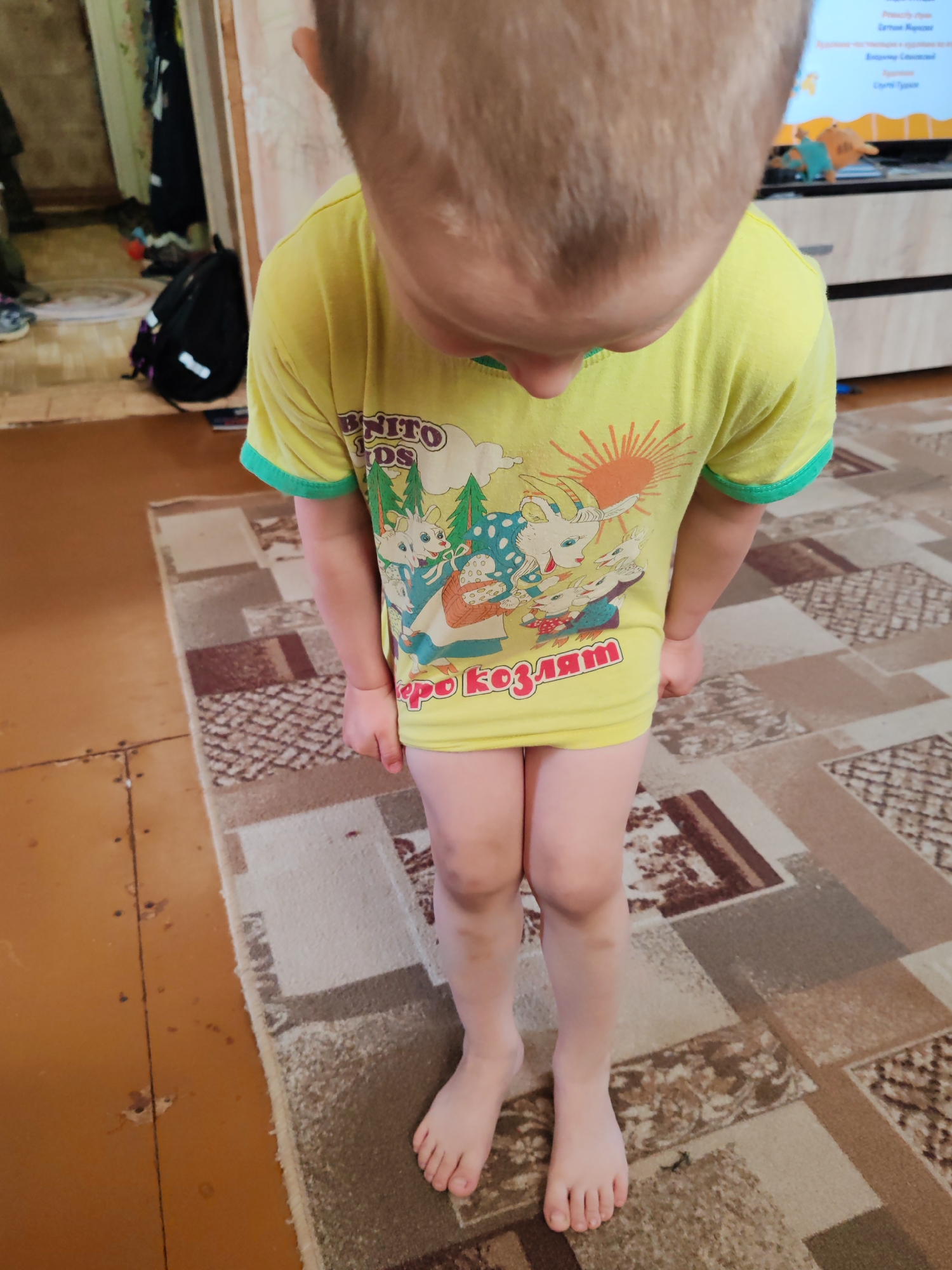 A child wearing a yellow t-shirt with a cartoon design is standing on a patterned rug. The rug is on a floor that appears to be wooden. The child's head is tilted downwards. A backpack and other items are visible in the background. The location is Чухлэм, Russia.
