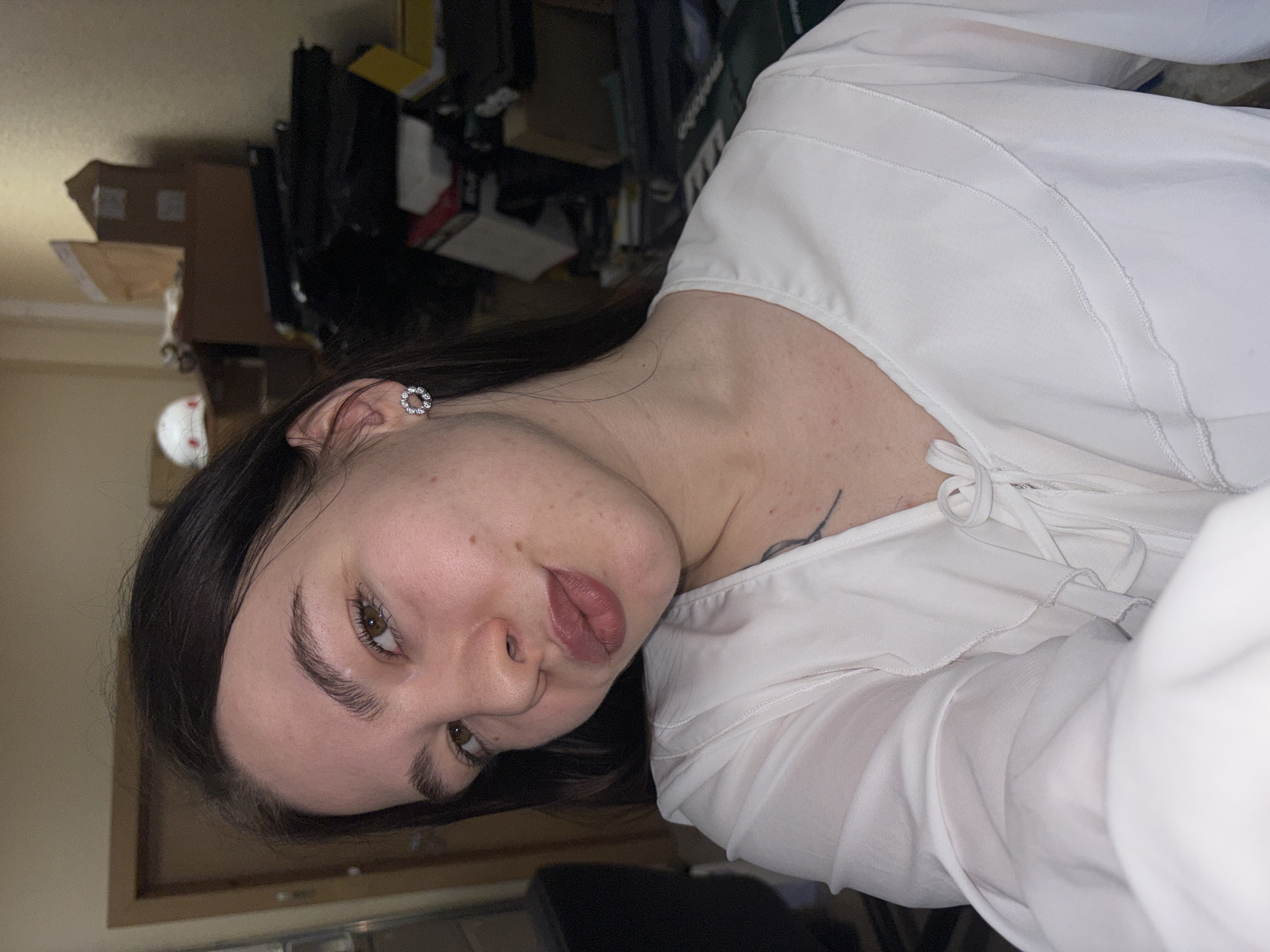 A woman with dark hair is facing the camera in an indoor setting. She has fair skin and is wearing a white blouse with tie details at the neckline. Her ear is visible, adorned with a round earring. A dark tattoo is partially visible on her upper chest. The background shows cluttered shelves with boxes and various objects. It is not possible to determine the location as the city of Perm, Russia based on this image alone.