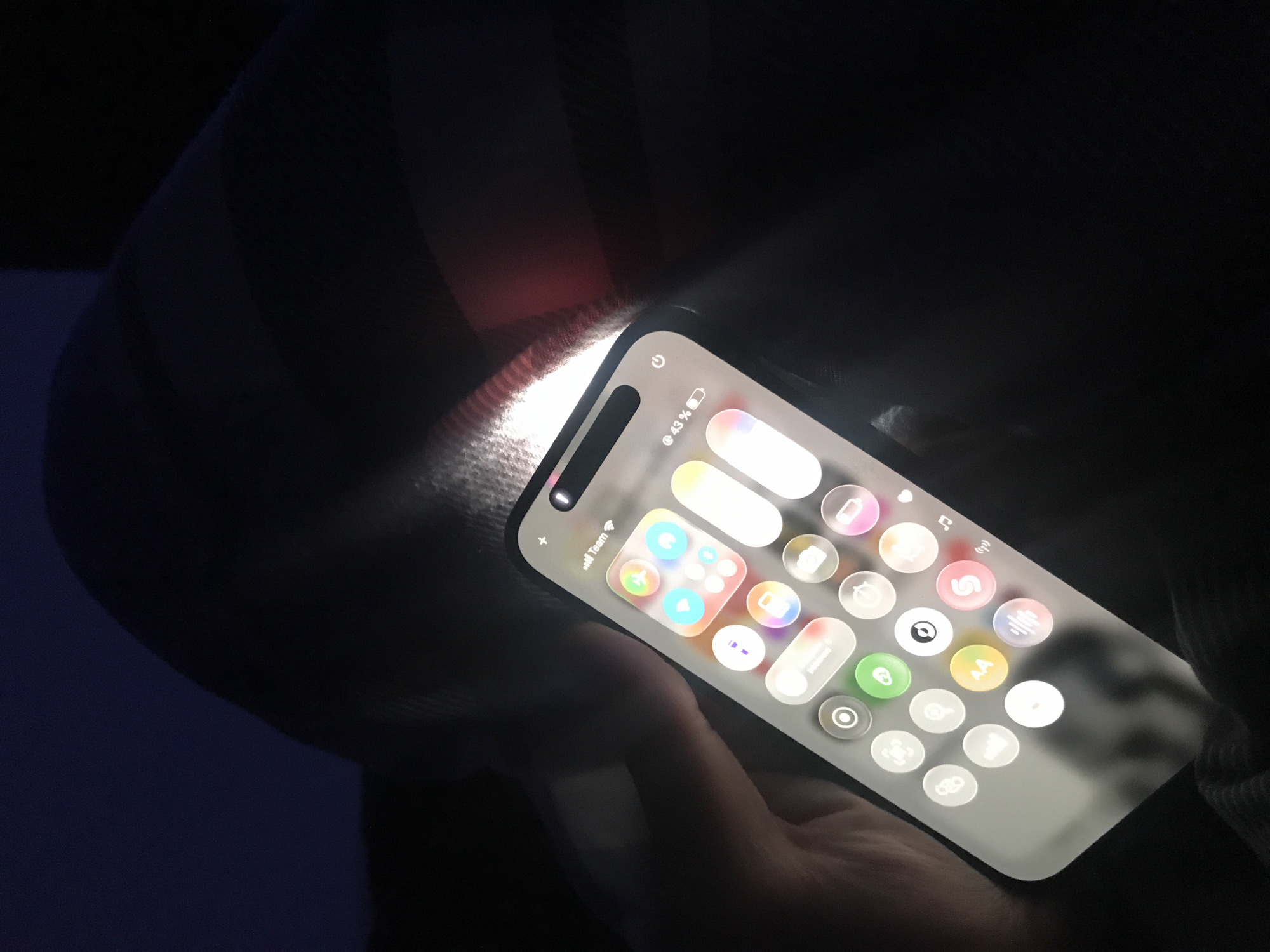 A person is holding an iPhone displaying the iOS Control Center in a dark environment. The screen is brightly illuminated, showing various control toggles and icons. The battery indicator shows 43% charge. The person's hand and clothing are partially visible. The location is unknown and there is no information indicating it is in Yerevan, Armenia.