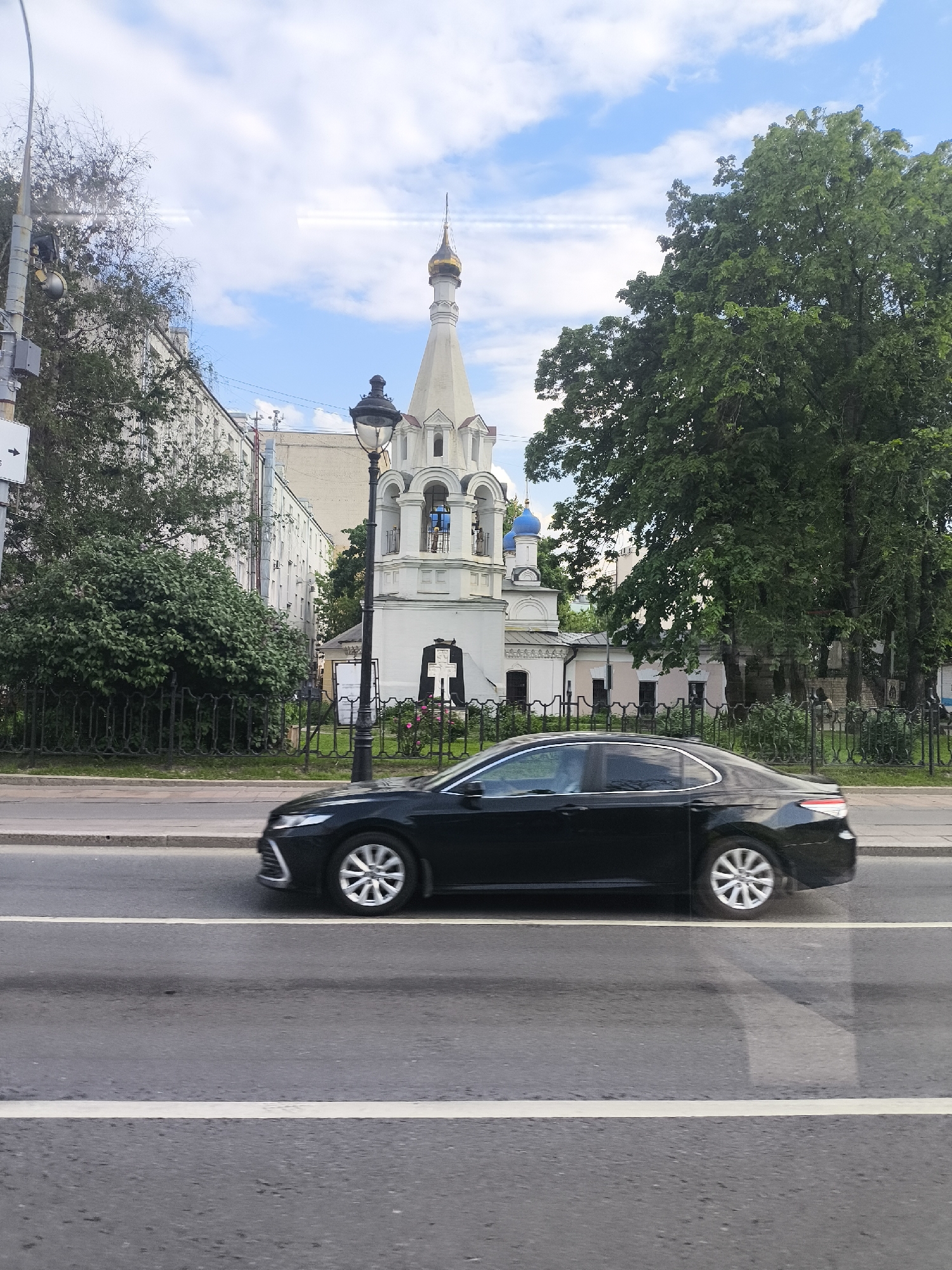 The image shows a black sedan moving on a multi-lane city street in an unknown location. The car is moving from left to right. Behind the car and across the street is a church with a white spire and golden dome, with blue onion domes visible on an adjacent structure. A black wrought iron fence borders the building. There are buildings and trees surrounding the church. A street lamp is visible near the fence. The sky is partially cloudy.