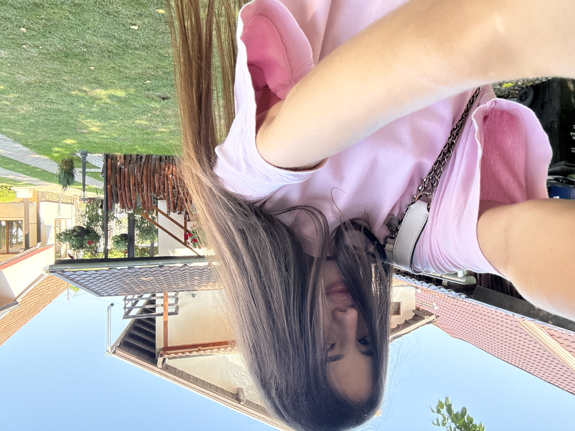 The media file is a close-up, selfie-style shot of a young woman with long, brown hair. She is wearing a pink t-shirt and a white and silver purse with a chain strap is visible. Her face is partially obscured by her hair and the angle of the shot, but she appears to be looking at the camera. The background suggests an outdoor setting, likely a backyard or garden area, with green grass and a paved walkway. In the distance, there are glimpses of houses with tiled roofs and some greenery. The lighting indicates it is daytime, likely late afternoon due to the warm tones. The overall mood is casual and relaxed. No visible text is present in the image. The location context is Cârțișoara, Romania.