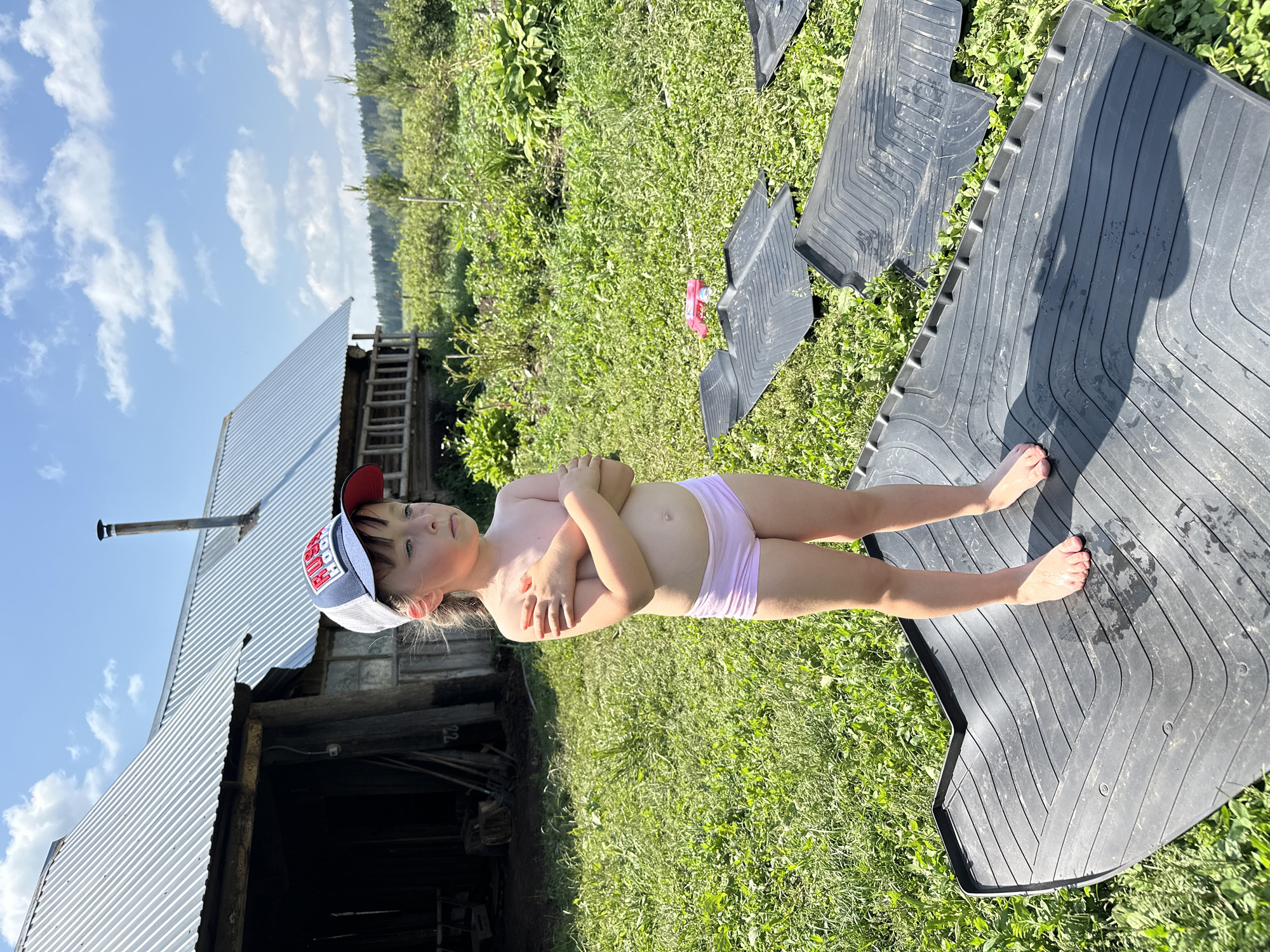A young girl, wearing a cap and pink underwear, stands with her arms crossed in a grassy yard. She is positioned in front of a rustic building with a corrugated metal roof and a chimney. The background features lush green foliage and trees, suggesting an outdoor, rural setting. In the foreground, several black rubber floor mats are laid out on the grass. The sky is visible above with scattered clouds, indicating it is daytime, likely a sunny day. The overall impression is one of a casual, everyday moment in a summery environment. The cap the girl is wearing has text and a logo on it.