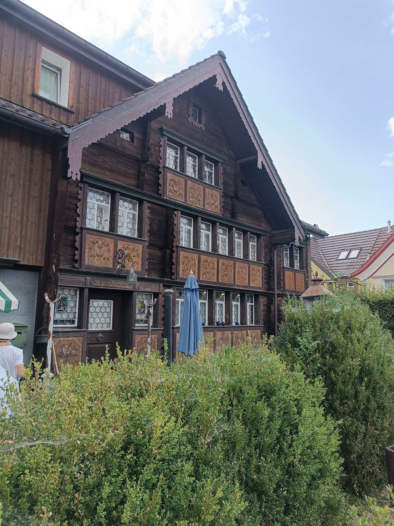 The image shows a traditional Swiss wooden house with ornate carvings and a steeply pitched roof. The house is located in the city of Appenzell, Switzerland, a region known for its well-preserved architecture. In the foreground, lush green bushes partially obscure the lower part of the house. A person wearing a white hat and shirt stands near the entrance of the house, partially visible from behind. A blue umbrella is also visible in front of the house. The sky above is a clear blue with scattered clouds. The building exhibits intricate decorative elements, including carved wooden panels featuring floral and grapevine motifs, and leaded glass windows with geometric patterns. The overall scene conveys a sense of traditional Swiss rural life and architecture.