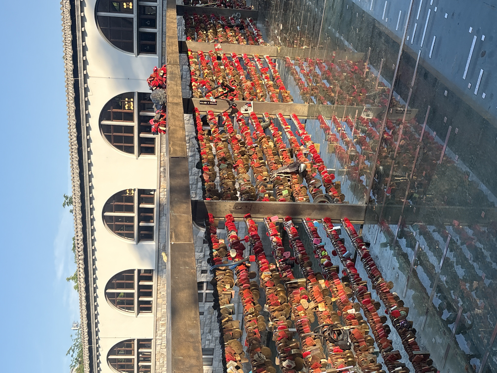This is a medium shot of a bridge covered in love locks, reflecting in the water below. The bridge is made of metal and wood, and the locks are of various colors, predominantly red, gold, and pink. Some locks have inscriptions on them. In the background, there is a white building with arched windows and a tiled roof. The sky is blue and clear. The overall scene is bright and appears to be bathed in sunlight.