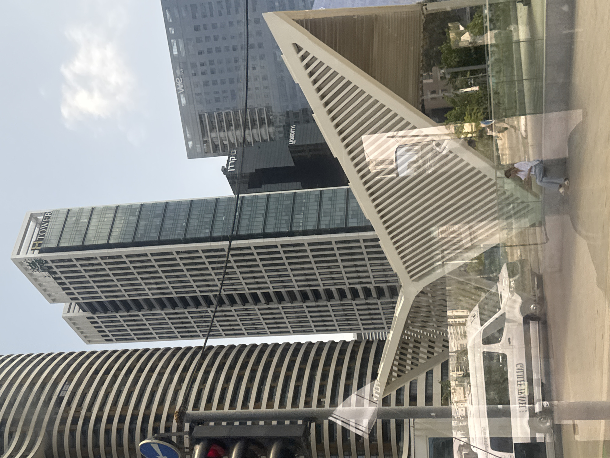 The image depicts a street scene in Tel Aviv, Israel, taken from behind a glass pane with reflections. Buildings of varying architectural styles dominate the view, including skyscrapers with grid-like facades and a curved building with striped horizontal lines. An abstract, triangular structure stands in the foreground, partially obstructing the view of the buildings. The reflections on the glass show a pedestrian sitting on a bench, a van with writing in Hebrew and English and greenery on the sidewalk. Street signs and a traffic light are also visible.