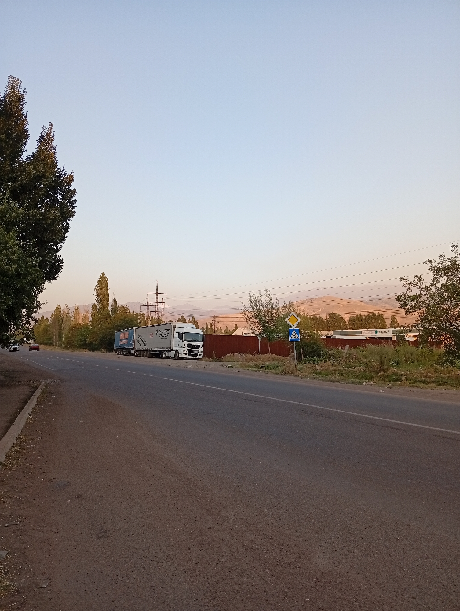 In Angren, Uzbekistan, a highway is visible with a white semi-trailer truck parked on the right side of the road. The truck has two trailers, one white and one blue. The white trailer displays "TARGOR TRUCK" and "SPECIAL TRANS." The blue trailer shows "GRAFS TRANSPORT." A red car is driving on the left side of the highway. A pedestrian crossing sign and a diamond-shaped warning sign are posted on the right side of the road. Tall trees line the left side of the highway, and in the distance, mountains are visible under a clear sky. The ground is paved with asphalt, and a concrete curb runs along the edge of the road.