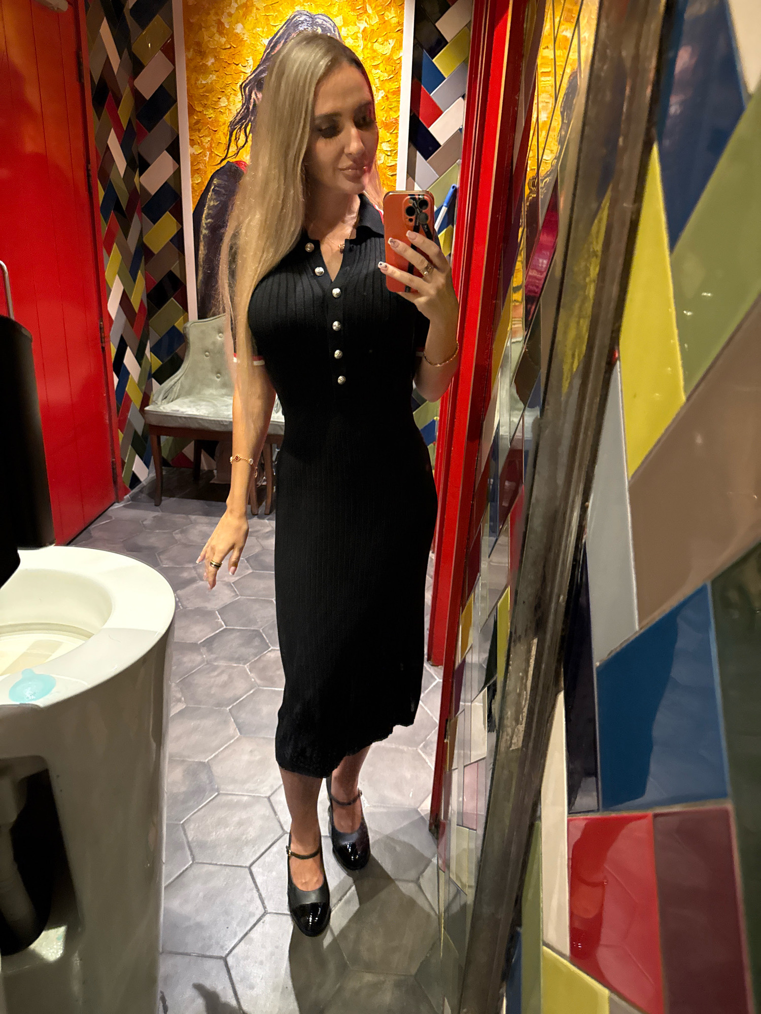 A woman with long blonde hair is standing in a bathroom, taking a selfie in a mirror. She is wearing a black, fitted, knee-length dress with a collared neckline and silver buttons. Her hair is partially covering her face as she looks down at her phone. She has on black Mary Jane style shoes. Next to her, to her left, is a white toilet. The bathroom features a brightly colored, mosaic tile wall with geometric patterns. A red door is visible to the far left of the image. In the background, a light green chair with upholstery is visible. The floor is tiled with grey hexagonal tiles. The image is taken from a low angle. This scene is not identifiable as being in Dubai, United Arab Emirates.