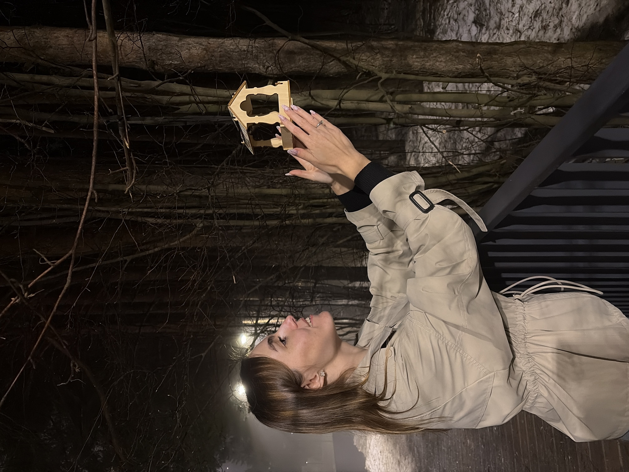 A woman stands outdoors in Izhevsk, Russia, holding a small, wooden birdhouse. The background features a dense collection of bare tree branches and trunks, possibly within a park or wooded area. The lighting is dim, suggesting it is either evening or a poorly lit area. The woman is wearing a light-colored trench coat with dark trim, and her hair is long and brown. She appears to be looking upwards, possibly at the birdhouse. There is a metal railing or structure visible, indicating she may be standing on a raised platform or path.