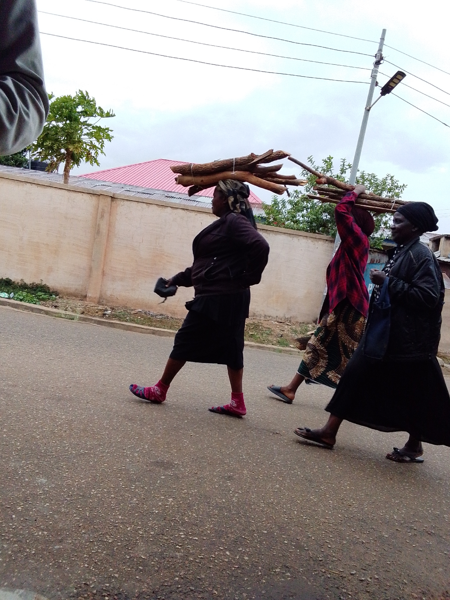 Three women are depicted walking along a paved road under an overcast sky, likely in Jos, Nigeria. The scene suggests a daytime setting, possibly a residential or semi-urban area.
The woman in the foreground on the left wears a dark top, a dark skirt, and striking pink and blue patterned socks, carrying a small dark object in her left hand. Next to her, a second woman carries a large bundle of firewood on her head, steadying it with her raised right arm. She is dressed in a red and black plaid long-sleeved shirt, a green and brown patterned skirt, and dark flip-flops. Further to the right, the third woman, wearing a dark top, dark skirt, and a dark head covering, also carries a bundle of sticks on her head and a blue bag over her left shoulder, walking in dark flip-flops.
Along the left side of the road, a light-colored wall or fence is visible, behind which a small green tree and a red corrugated roof can be seen. On the right, a utility pole with an LED street light and overhead power lines extends into the cloudy sky. The women are engaged in the activity of transporting firewood, moving from left to right across the frame. Their expressions are generally neutral or slightly smiling, as seen on the woman on the far right. No visible text is present in the image.
