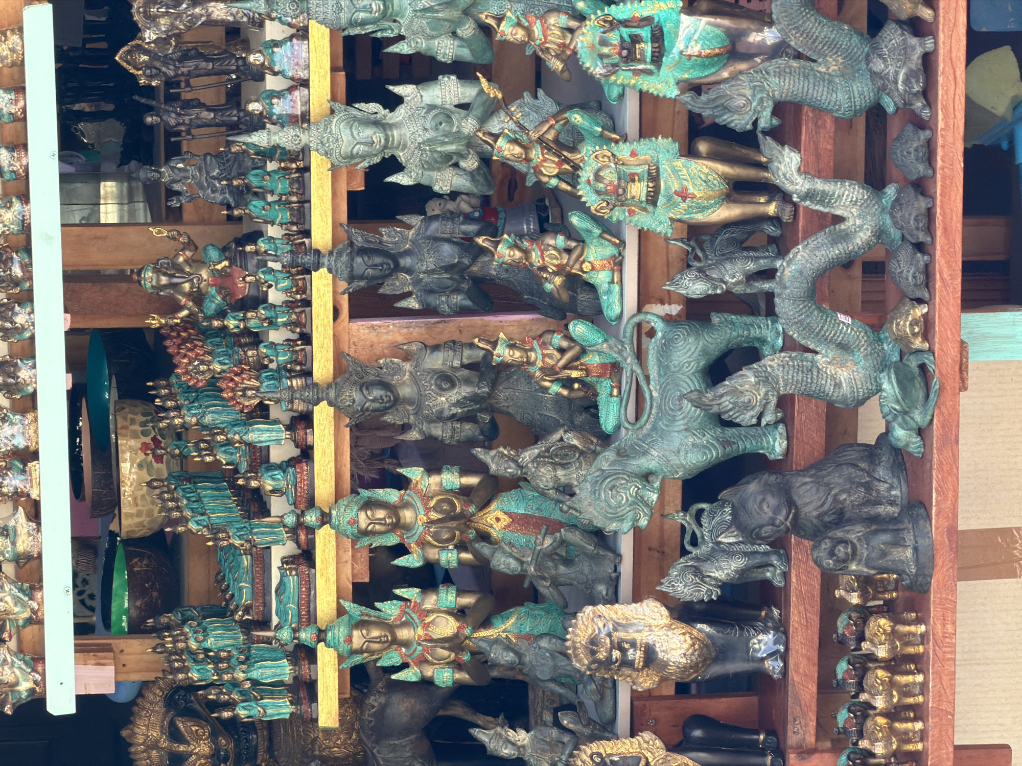 The image shows a market stall in Baan Plai Laem, Thailand, filled with various bronze and gilded statuettes and figurines. The items are displayed on wooden shelves. The subjects include numerous small figures of deities, possibly Buddhist or Hindu, with some featuring elaborate headdresses and flowing robes. There are also animal figurines, such as lions with golden manes, elephants, a cat, a dog, a horse, a dragon-like creature with multiple heads, and turtles. Some of the human figures are depicted in prayer or playing musical instruments. The color palette is dominated by bronze and teal patinas, with accents of gold and red. The lighting suggests an outdoor or semi-enclosed market setting, with soft shadows and a warm overall tone. There are no people clearly visible in the image.