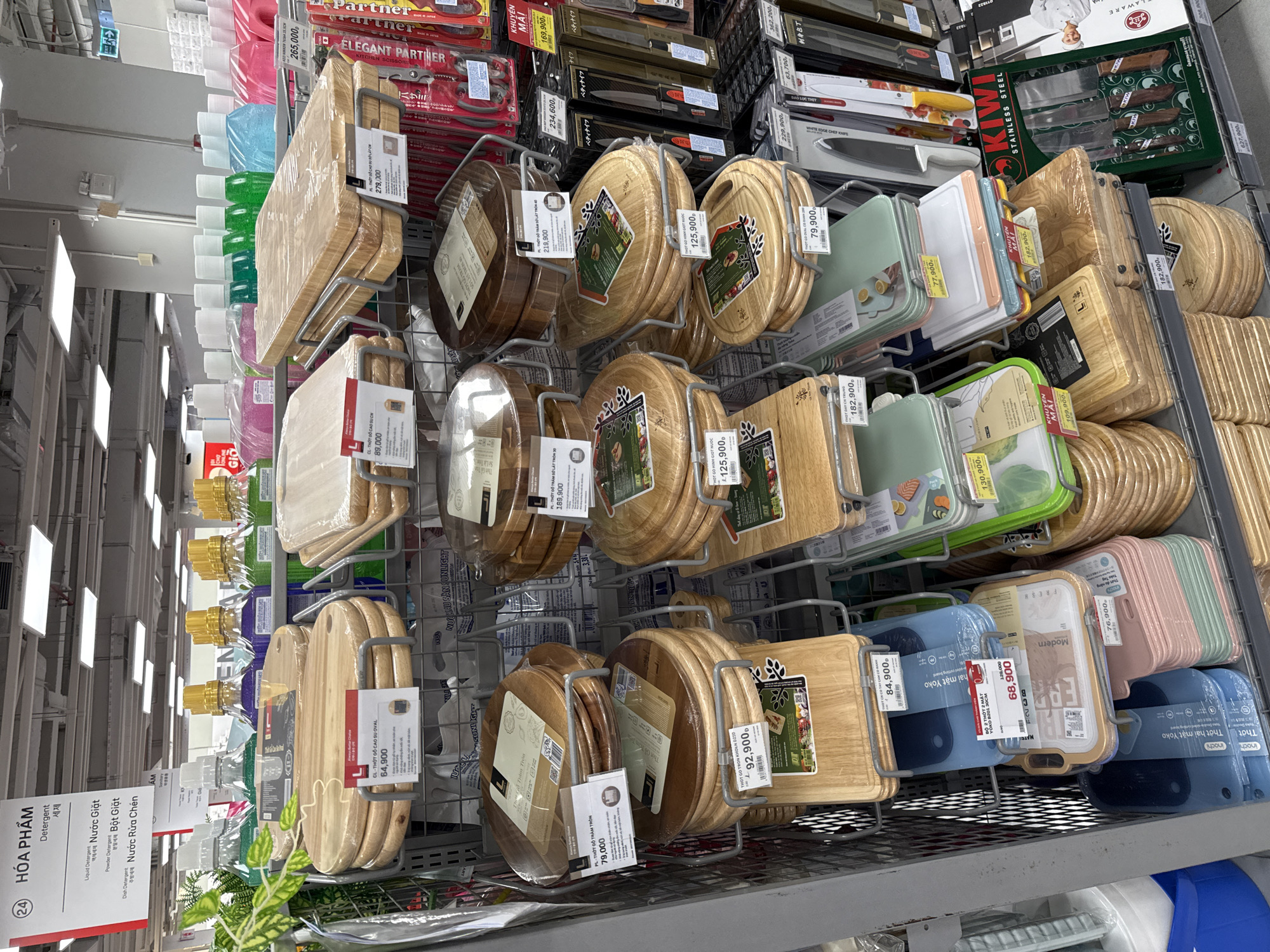 The image depicts a retail store, likely a supermarket or department store, in Nha Trang, Vietnam, showcasing a variety of kitchenware, primarily cutting boards. The items are neatly organized on metal shelves and displays. The cutting boards are made of wood and plastic, in various shapes and sizes, with price tags attached. In the background, detergent bottles and other household items are visible on shelves. The lighting suggests an indoor environment, typical of a commercial establishment. There are no people visible in the image. The overall scene is one of commercial retail display of kitchen products.