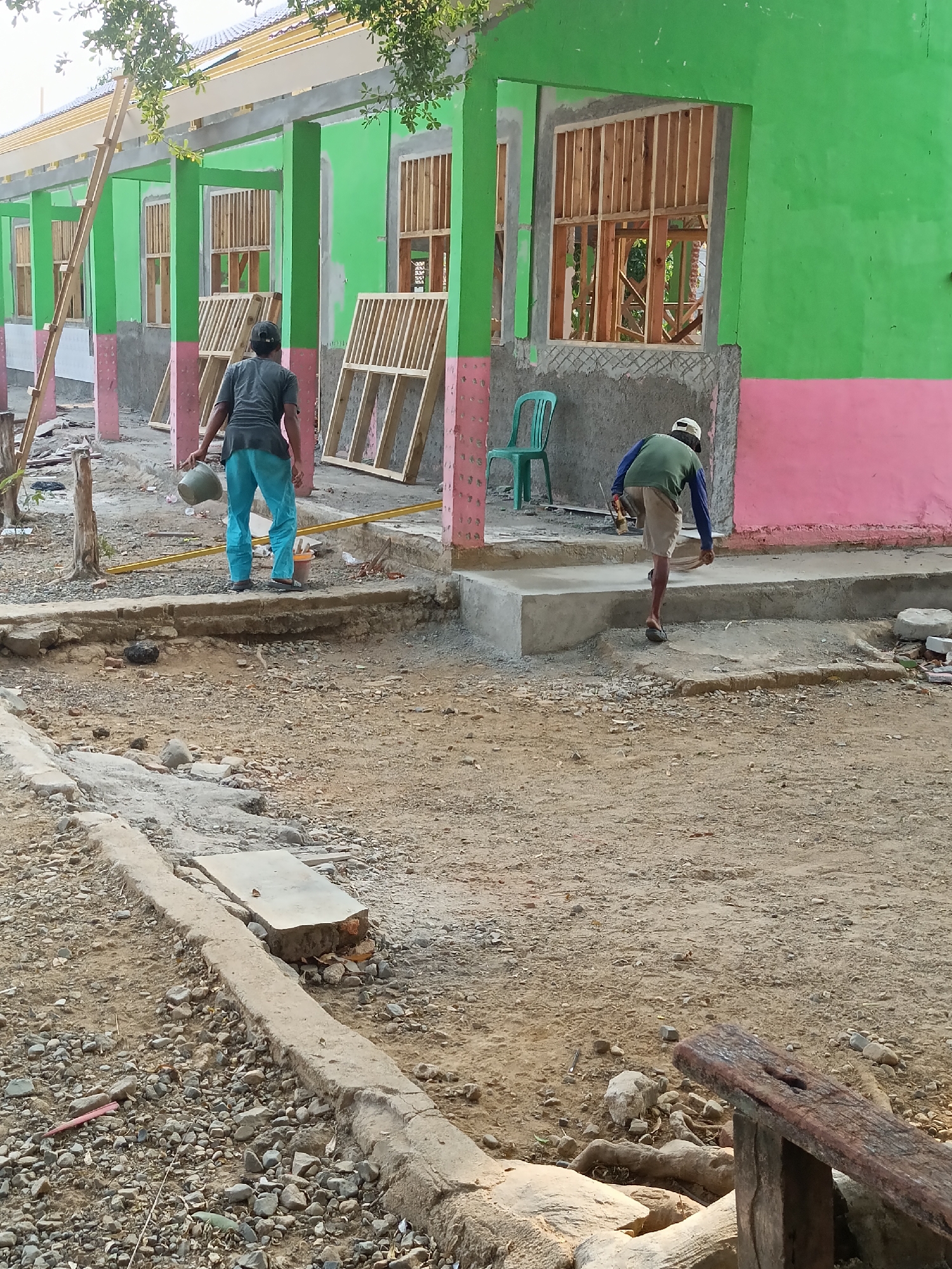 The image depicts a construction site in Sumberejo, Indonesia, likely a school or public building, given the row of unfinished rooms with green and pink exteriors. Two men are present, engaged in construction activities. One man, wearing a dark t-shirt and light blue pants, carries a grey bucket. The other man, in a green long-sleeved shirt and beige shorts, is seen walking up a concrete staircase, holding a trowel and what appears to be a bag of cement. The environment is dusty and filled with construction debris, including wooden framing, concrete slabs, rocks, and scattered tools. The building itself shows unfinished walls and window frames, indicating ongoing renovation or construction. The overall scene suggests a typical day at a construction site in a rural or semi-rural Indonesian setting.