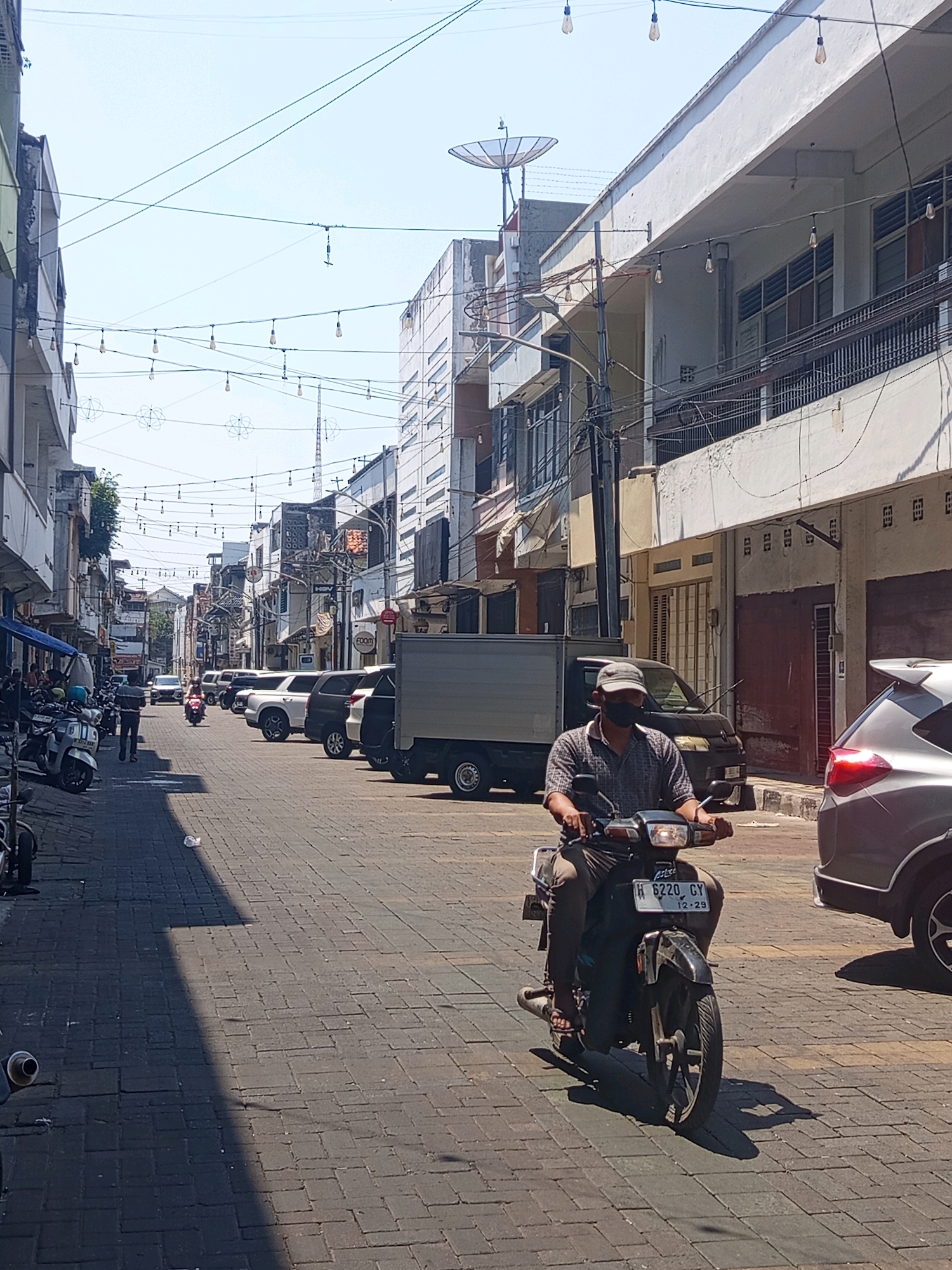 The image depicts a street scene in Semarang, Indonesia, characterized by a row of buildings on either side of a paved road. The architecture suggests an urban environment with a mix of commercial and residential structures. Overhead, a series of string lights are visible, adding a decorative element to the street. On the right side of the road, a silver car is parked, and to its left, a man wearing a cap and a patterned shirt is riding a motorcycle. The motorcycle has a license plate that reads "H 6220 GY" and "12-29". The man is wearing a face mask. The road is paved with bricks, and shadows from the buildings and the overhead lights stretch across it. Several other vehicles, including cars and motorcycles, are parked along the left side of the street. The sky is clear and bright, indicating a sunny day. The overall impression is a bustling urban street with typical elements of city life in Indonesia.