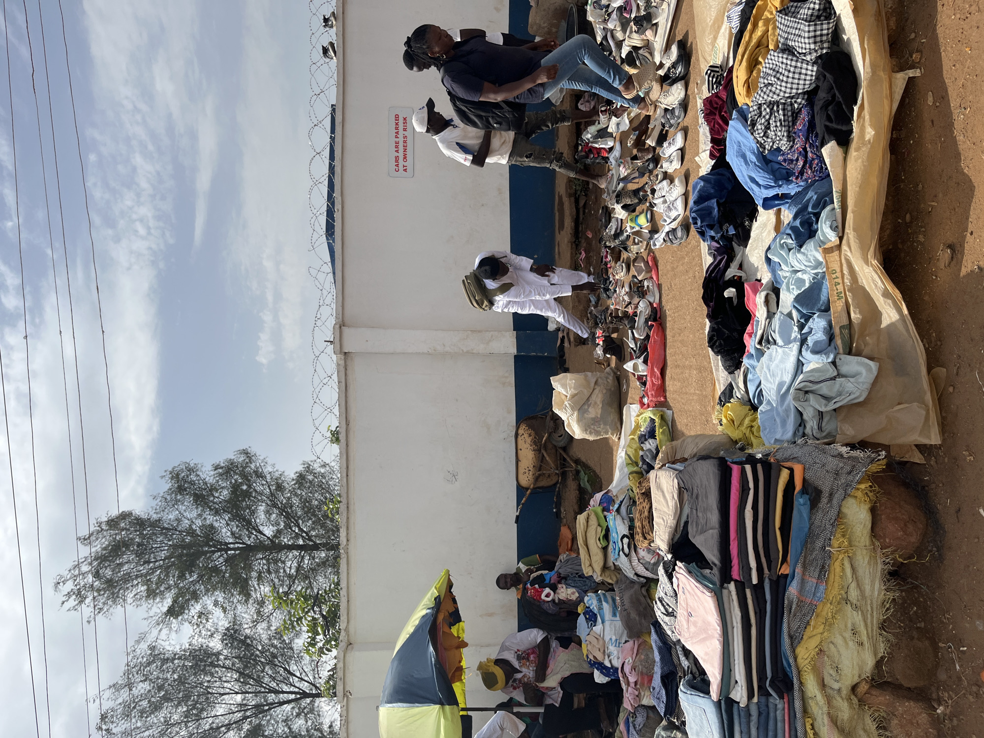 The image depicts an outdoor market scene in Rafin Par, Nigeria. In the foreground, an assortment of clothing items are spread out on tarpaulins, including stacks of folded shirts and pants, as well as loosely draped garments. Several individuals are present in the middle ground and background. One person wearing white is bent over examining shoes on the ground, while others, including a person in a navy shirt and blue jeans, walk through the market. A yellow and green umbrella provides shade for a vendor and a customer seated behind a display of clothes. A rusty wheelbarrow is visible against a white wall adorned with a sign that reads "CARS ARE PARKED AT OWNER'S RISK." Numerous pairs of shoes are laid out for sale in front of the wall. The environment appears to be a dusty, outdoor area with trees and a cloudy sky in the background.