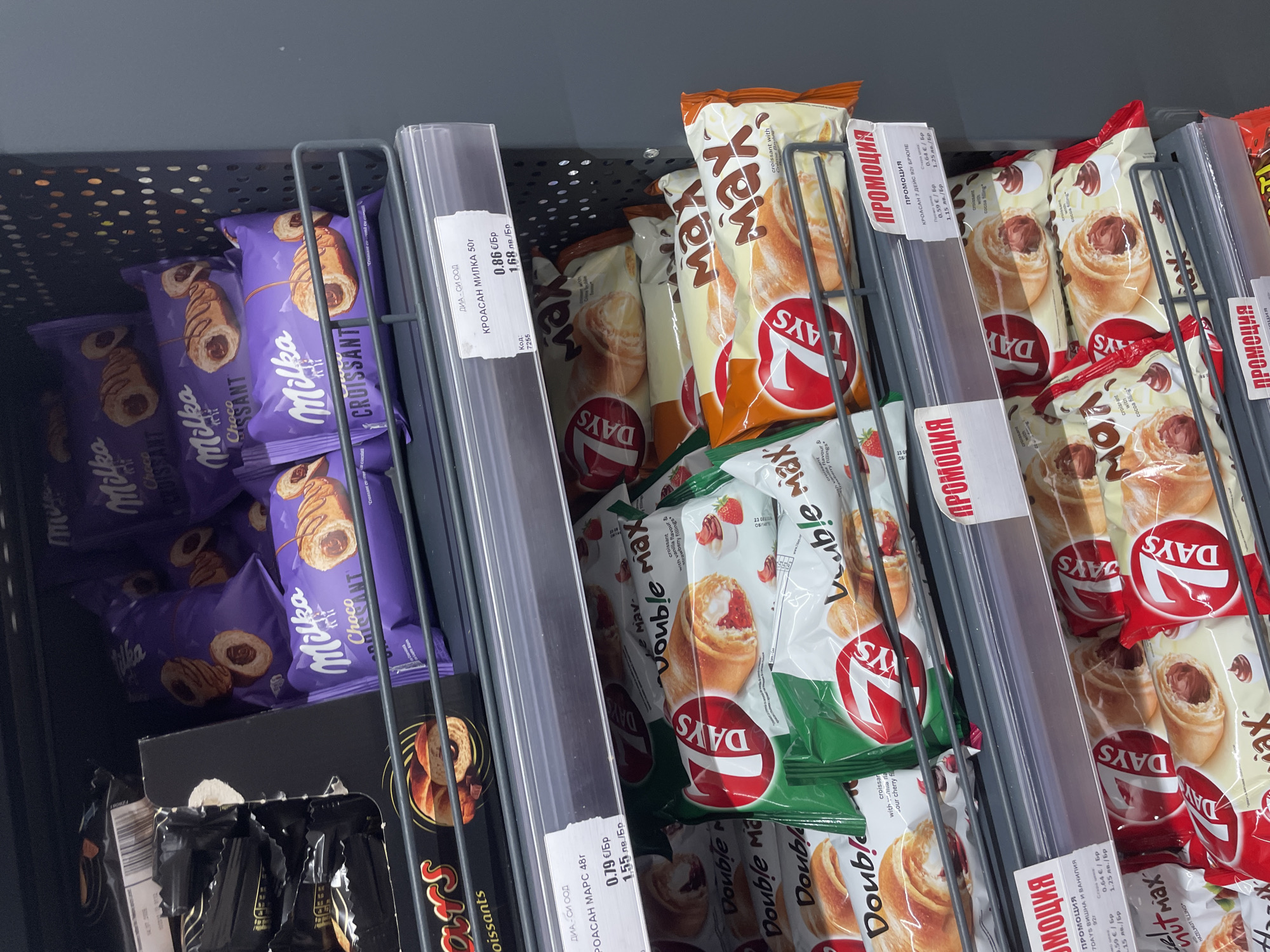 The image captures a top-down view of a store shelf displaying various packaged croissants, likely in a supermarket or convenience store in Ihtiman, Bulgaria. The products are arranged in vertical racks.
The primary subjects are rows of Milka Choco Croissants in distinctive purple packaging, and multiple varieties of 7DAYS brand croissants. The Milka croissants feature a chocolate filling and are visible on the left side of the frame. To their right, 7DAYS croissants are prominent, including "Max" and "Double Max" versions. Flavors visible include hazelnut filling, cocoa filling, and varieties with strawberry filling and vanilla & sour cherry filling, indicated by their packaging designs (orange for hazelnut/cocoa, green for strawberry, and white/red for cherry/vanilla).
The activity taking place is product display for retail sale. Several price tags are visible, written in Bulgarian. One tag for "КРОАСАН МИЛКА 50г" (Milka Croissant 50g) shows a price of 0.86 €/Br (BGN) or 1.68 лв./Br (BGN). Another tag mentions "КРОАСАН МАРС 48г" (Croissant Mars 48g) at 0.79 €/Br or 1.55 лв./Br, though the visible product next to it is a 7DAYS croissant. Multiple "ПРОМОЦИЯ" (PROMOTION) tags are present for 7DAYS products, detailing old and new prices in both Euro and Bulgarian lev per piece, such as 0.59 €/Br or 1.15 лв./Br from a previous 0.64 €/Br or 1.25 лв./Br. The text "ДИА - СИ ООД" (DIA - SI OOD), likely a store or distributor name, also appears on some tags. The store environment is generic with black perforated shelving and bright, artificial lighting, offering no specific time of day or weather cues.