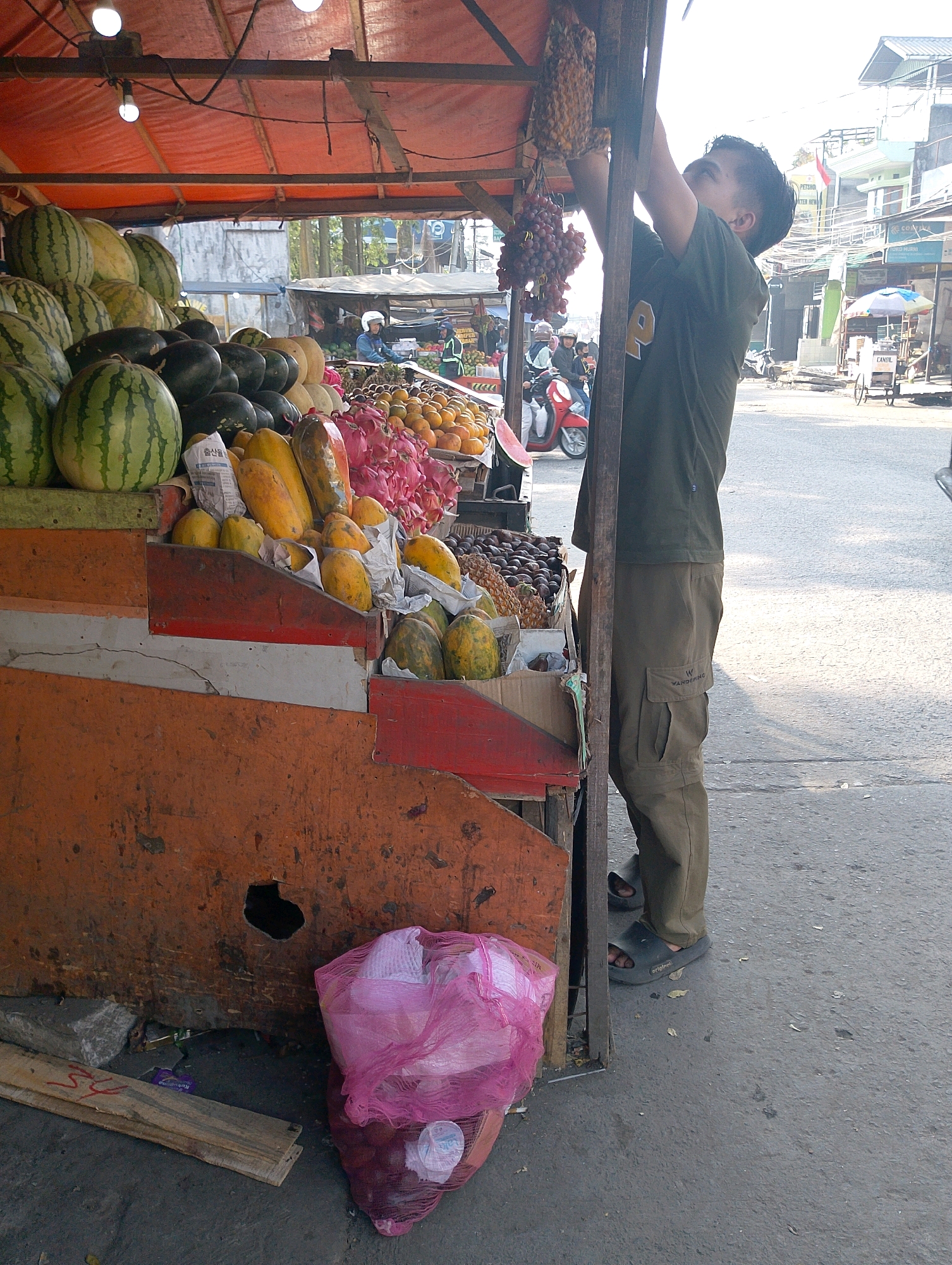 In Lembang, Indonesia, a young man in a dark green t-shirt and khaki pants is seen reaching up to adjust a bunch of grapes hanging from a wooden support of a fruit stall. The stall is covered with an orange tarp, and brightly lit by two bare light bulbs. In front of the stall, various fruits are displayed on tiered wooden shelves, including watermelons, papayas, pineapples, and dragon fruit. The ground is paved with asphalt, and in the background, there are buildings and other market stalls. A pink mesh bag filled with produce sits on the ground to the right of the stall. The scene depicts a typical street market environment in Lembang.