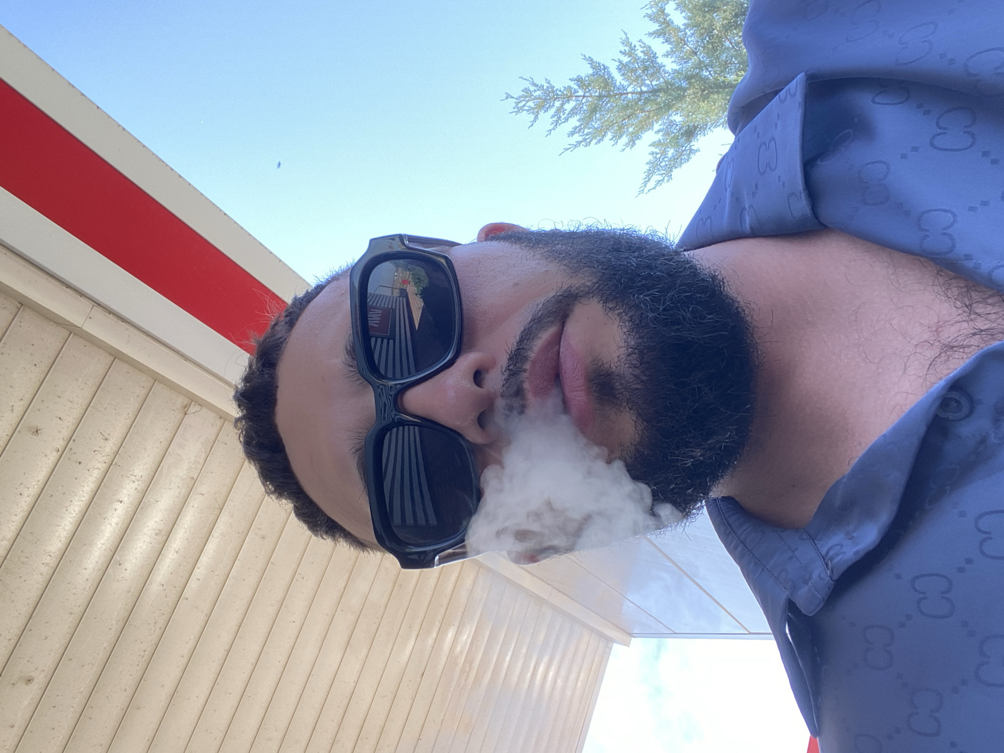 A low-angle, medium shot captures a man exhaling smoke while looking up at the sky. He is wearing black sunglasses and a dark blue shirt with a repeating Gucci logo pattern. Smoke emanates from his mouth and obscures part of his face. The background features a wooden ceiling and a clear blue sky with a thin tree on the right. The image appears to have been taken outdoors, possibly at a gas station, given the structure of the ceiling and the bright lighting. The specific location in Korkmaskala, Russia, is not discernible from the visual cues in the image.