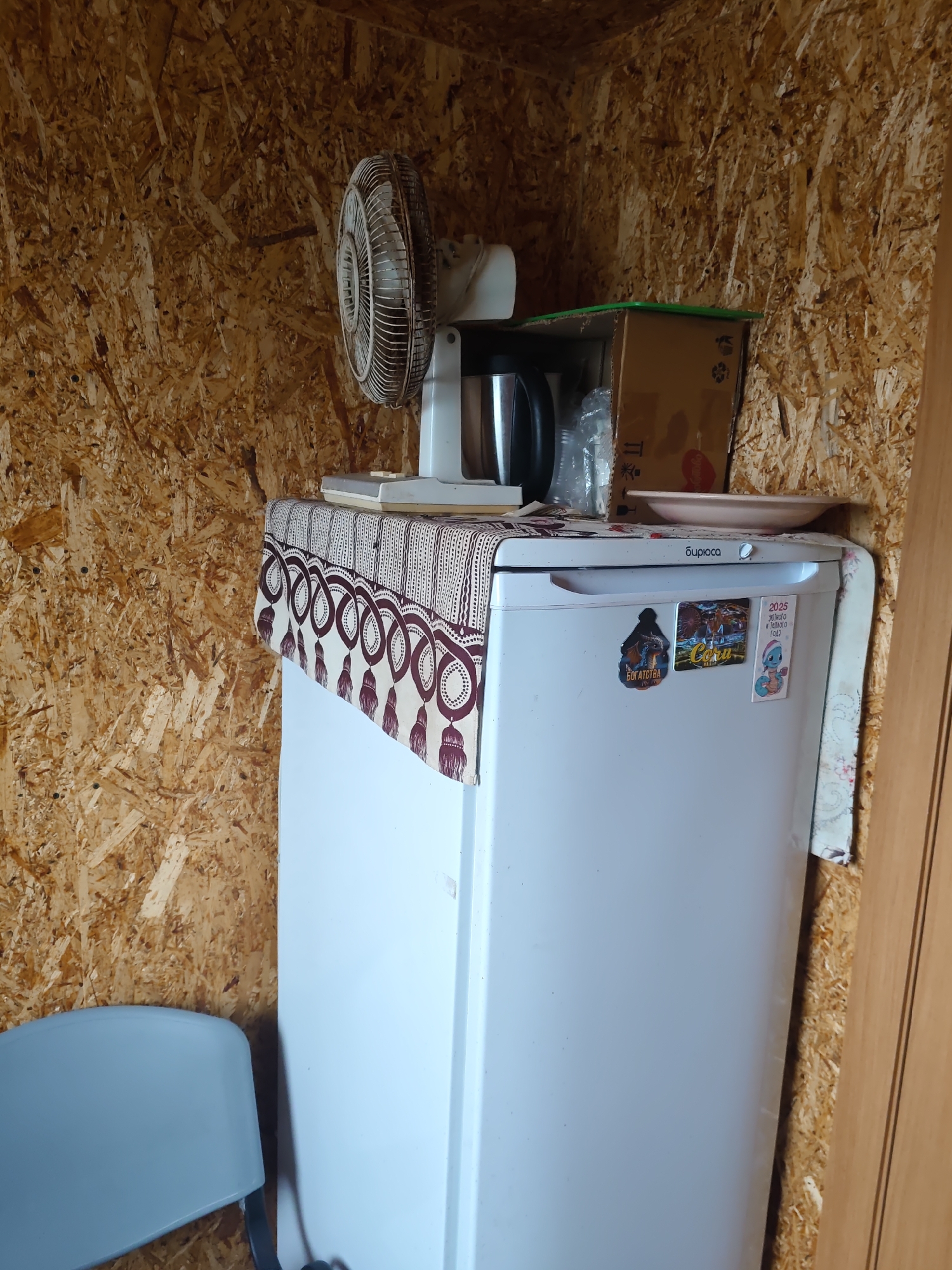 The image shows an interior scene within a structure with walls made of oriented strand board (OSB). In the center is a white refrigerator, likely a compact model. On top of the refrigerator, a patterned tablecloth is spread out, covering some items including what appears to be a metal thermos or kettle, a box, and a light pink plate. To the left of the refrigerator, a white fan with a metal grill is positioned on top of the tablecloth. A light blue chair is partially visible in the lower left corner of the frame. The overall impression is that of a utilitarian space, possibly a shed, garage, or a small dwelling. No people are visible in the image. The specific location in the city of Платошино, Russia, cannot be determined from the visual information alone.