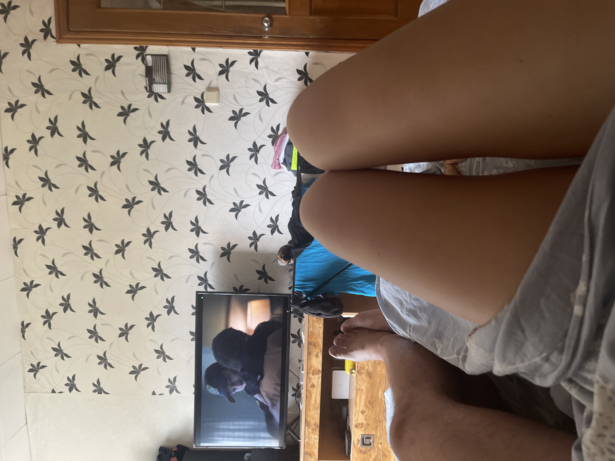 The image shows a close-up view of two pairs of legs lying on a bed, with a television and a dresser in the background. The room is decorated with floral wallpaper and has a wooden door. The scene appears to be a casual domestic setting. The location is identified as Shcherbinka, Russia.