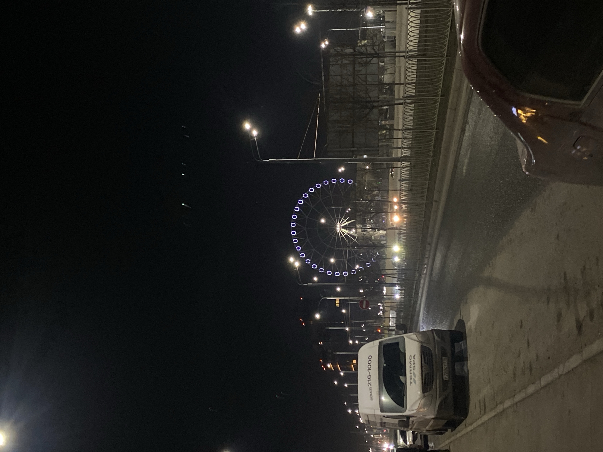 At night, the image shows a street in Kazan, Russia, with a lit Ferris wheel in the background. Street lights illuminate the road, and a white van with visible company information is parked on the side of the road. A car is visible in the upper right corner of the frame. The scene is urban, capturing city life at night.