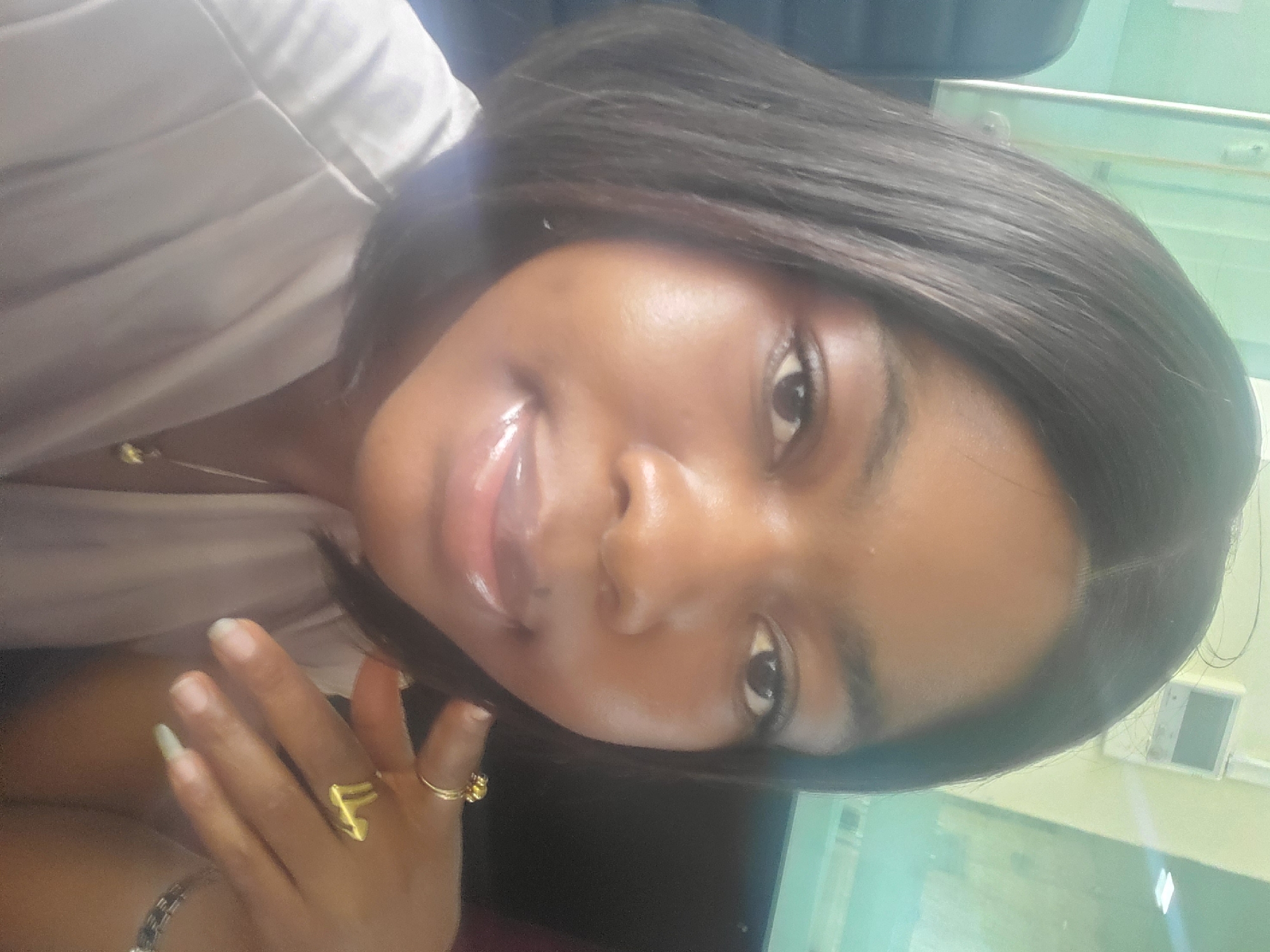 A close-up selfie of a woman with dark hair wearing gold rings and a light-colored shirt. She is likely indoors in Gwagwada, Nigeria, with an air conditioning unit visible in the background.