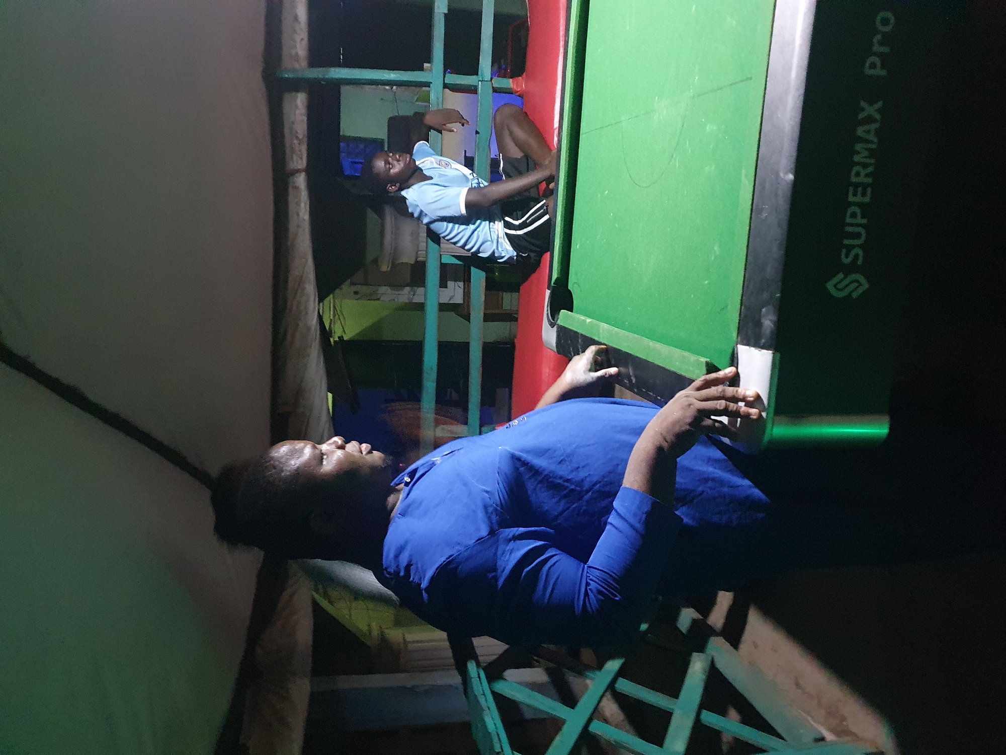 Two young men are present at an outdoor billiards table at night. The man in the foreground is standing and wearing a blue long-sleeved shirt and dark pants. The man in the background is sitting and wearing a light blue t-shirt with white stripes on the sleeves and dark shorts. The billiards table is green, and the area appears to be a recreational spot. There is a green railing in the foreground and a tent-like structure overhead. The setting is likely an informal gathering place.