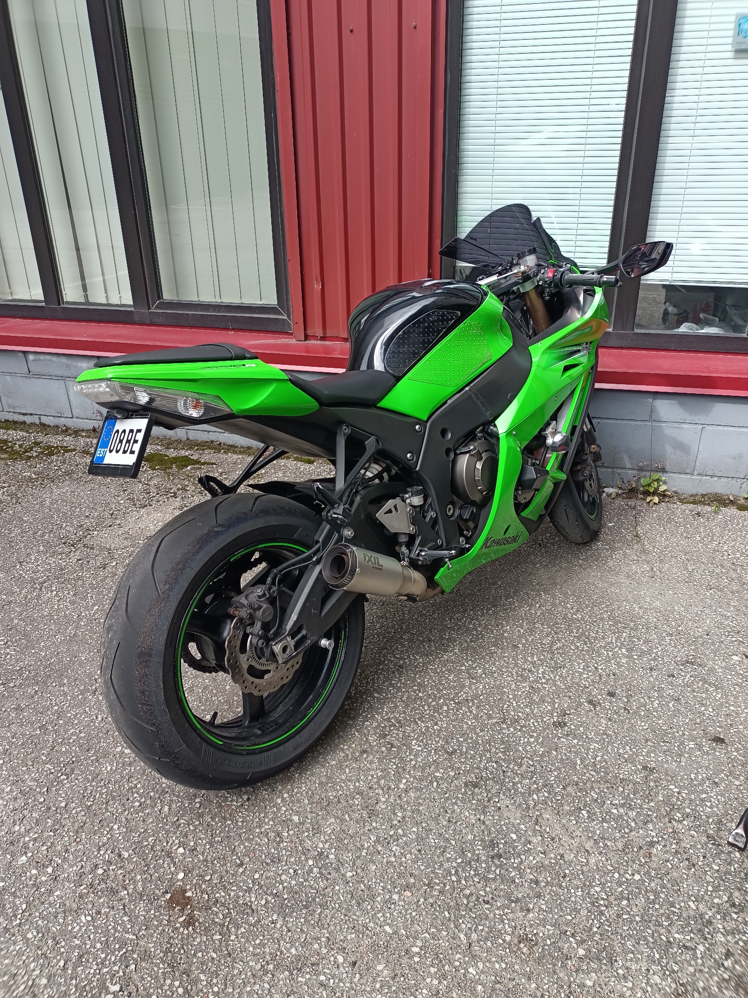 A green Kawasaki motorcycle is parked on asphalt in front of a building with a red metal facade and large windows with white blinds. The motorcycle is positioned at an angle, with its rear wheel closer to the camera. The license plate, which reads "08BE EST", is visible on the rear fender. The motorcycle has a black seat and tank, and prominent green bodywork. The building appears to be a commercial or industrial structure.