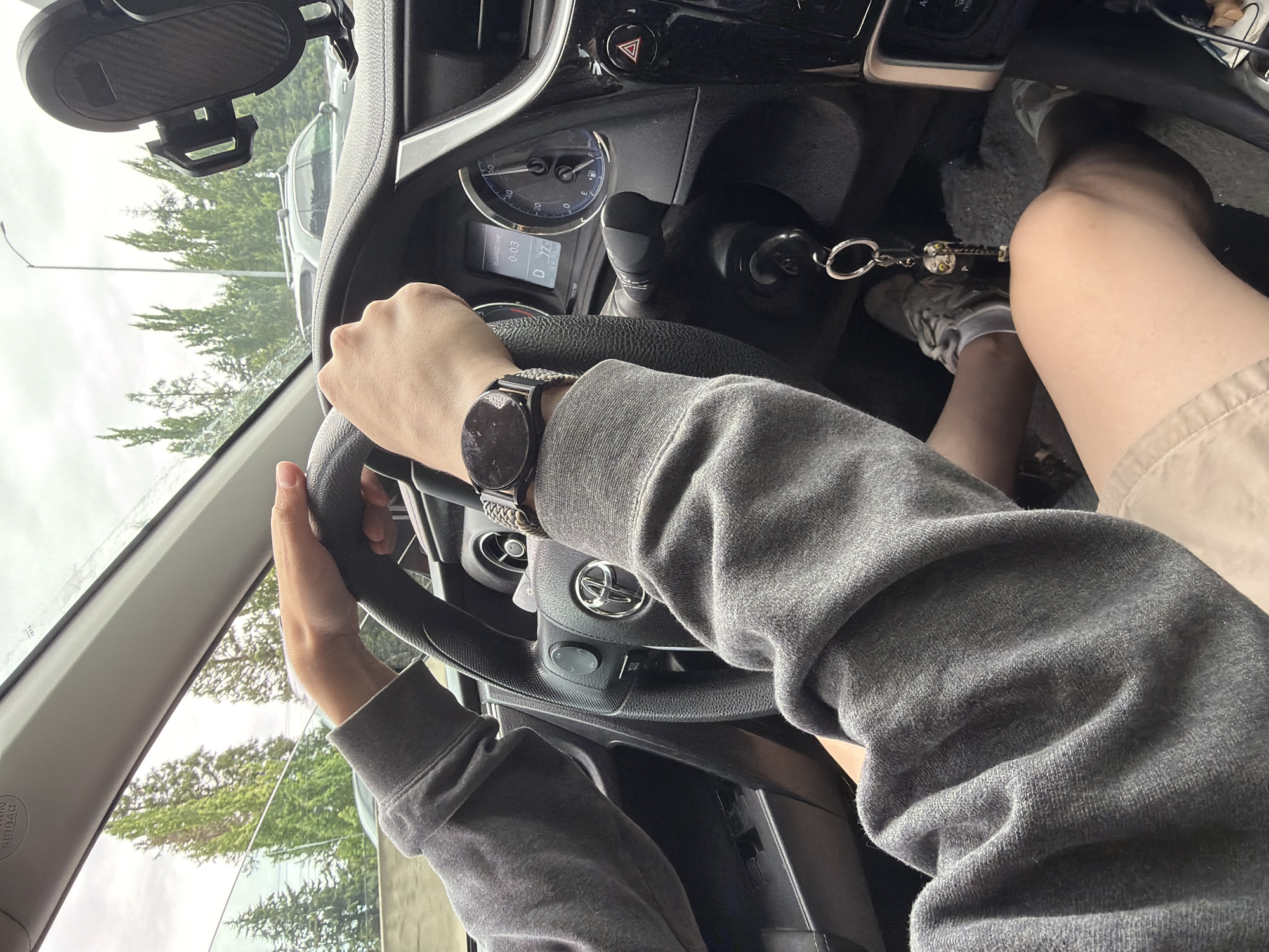 The image shows a first-person perspective from inside a car, with the driver's hands on the steering wheel. The driver is wearing a grey sweatshirt and a watch on their left wrist. Their legs are visible, wearing beige shorts and grey sneakers. A keychain with an owl charm is attached to the car's ignition.
The car's interior is visible, including the steering wheel with a Toyota logo, dashboard with gauges, and a gear shift. Outside the car, through the windshield and side windows, trees and an overcast sky are visible, suggesting it is daytime and potentially rainy or cloudy. A street lamp is also visible through the windshield.
The scene is set in Beaverton, USA. The driver appears to be either about to start driving or is stopped, as indicated by the gear shift position and the fact the car is not in motion. There is no clear indication of emotion.
Text visible includes "AIRBAG" on the car's interior pillar and numbers on the dashboard display that appear to be "003" and "D" suggesting the car is in drive.