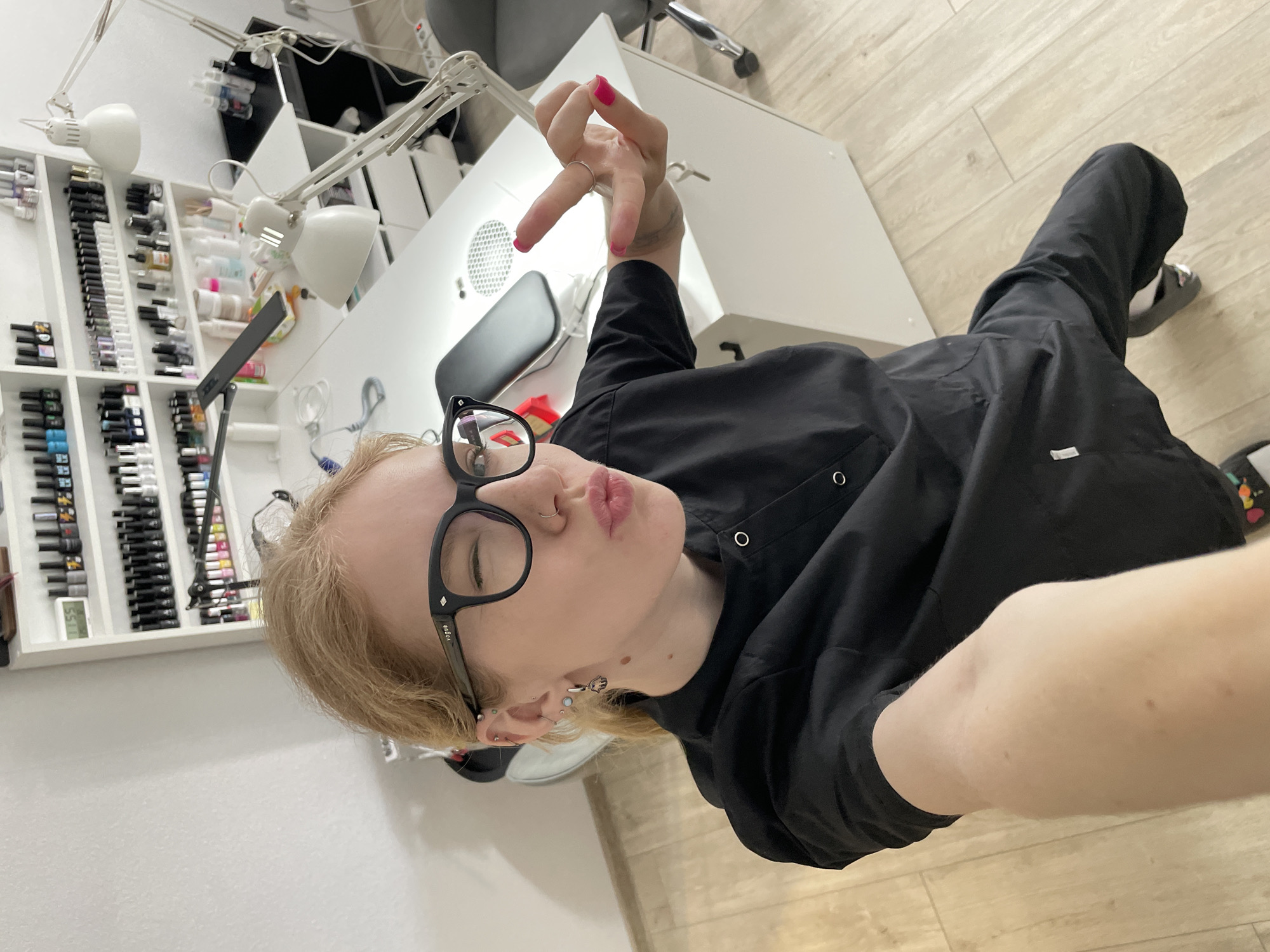 A light-skinned person with blonde hair, wearing black-framed glasses, a nose ring, and multiple earrings, is taking a selfie. They are wearing a black button-down garment. The person is making a kissing face and holding up their right hand with pink nail polish. The background shows a white desk with a vent, two white lamps, and a white shelf containing numerous nail polish bottles. The floor is light-colored wood. Based on the presence of nail polish and manicure tools, the scene appears to be a nail salon. There is no definitive information to conclude that this photo was taken in Izhevsk, Russia.