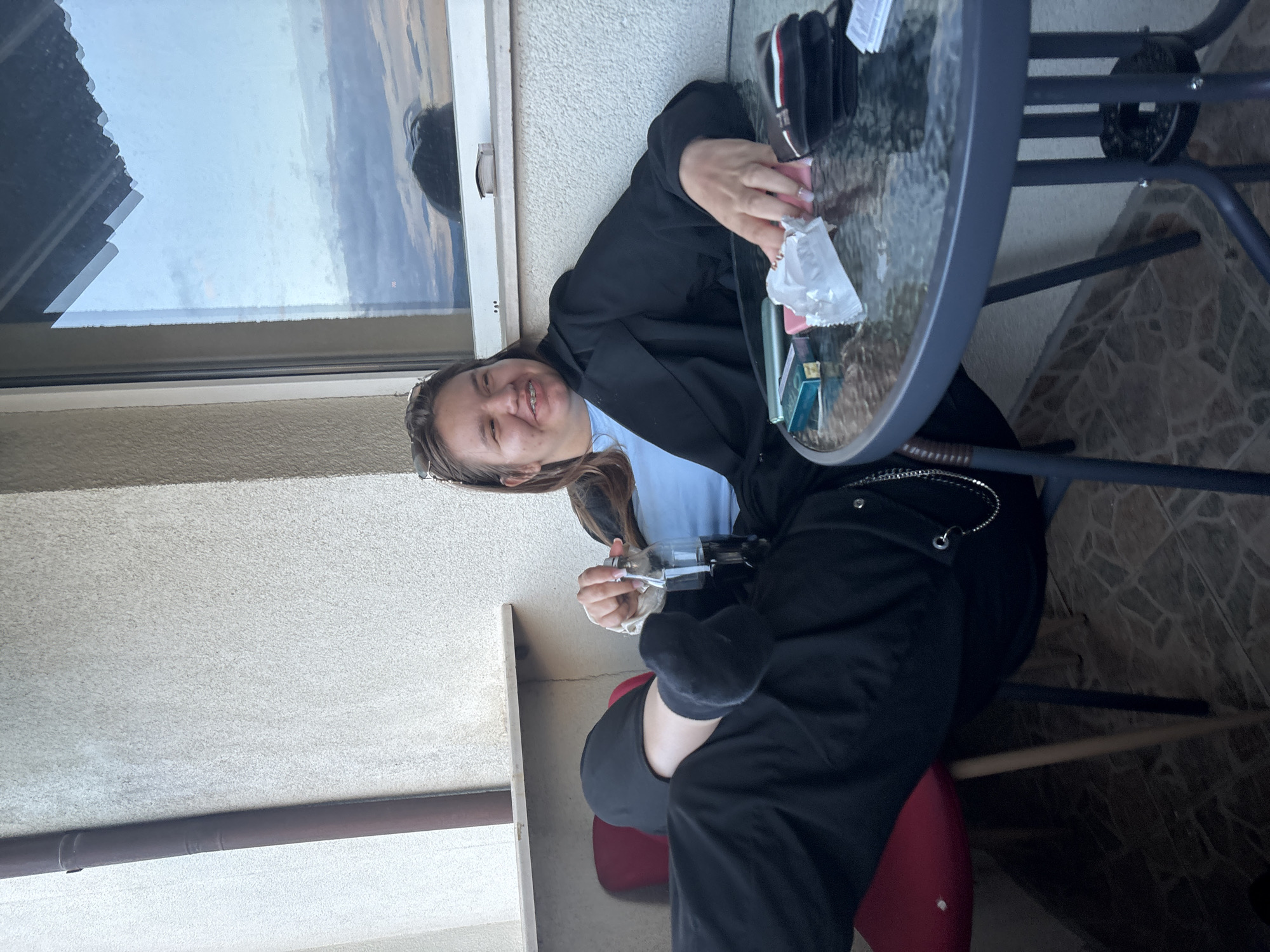 A young person with long brown hair, wearing a light blue shirt under a dark jacket, is seated at a small round table on an outdoor balcony. They are smiling broadly at the camera, revealing braces. In their hands, they hold a clear glass bottle with a stopper.
The table features a glass top and a dark metal frame. On the table, several items are visible, including a dark wallet or case with "TOMMY HILFIGER" faintly inscribed, a pink object, a crumpled piece of white paper or plastic, and a couple of blue/green rectangular boxes, one of which appears to be a "PARLIAMENT" cigarette pack.
The setting is a balcony with a light-colored wall. To the left, a window reflects an overcast or partly cloudy sky, suggesting it is daytime. Below the window, a dark horizontal pipe or railing is visible. The floor is tiled with a geometric pattern. The person is seated on a red chair, partially visible beneath the table. The overall scene suggests a casual, relaxed moment on a balcony in Blagoevgrad, Bulgaria.