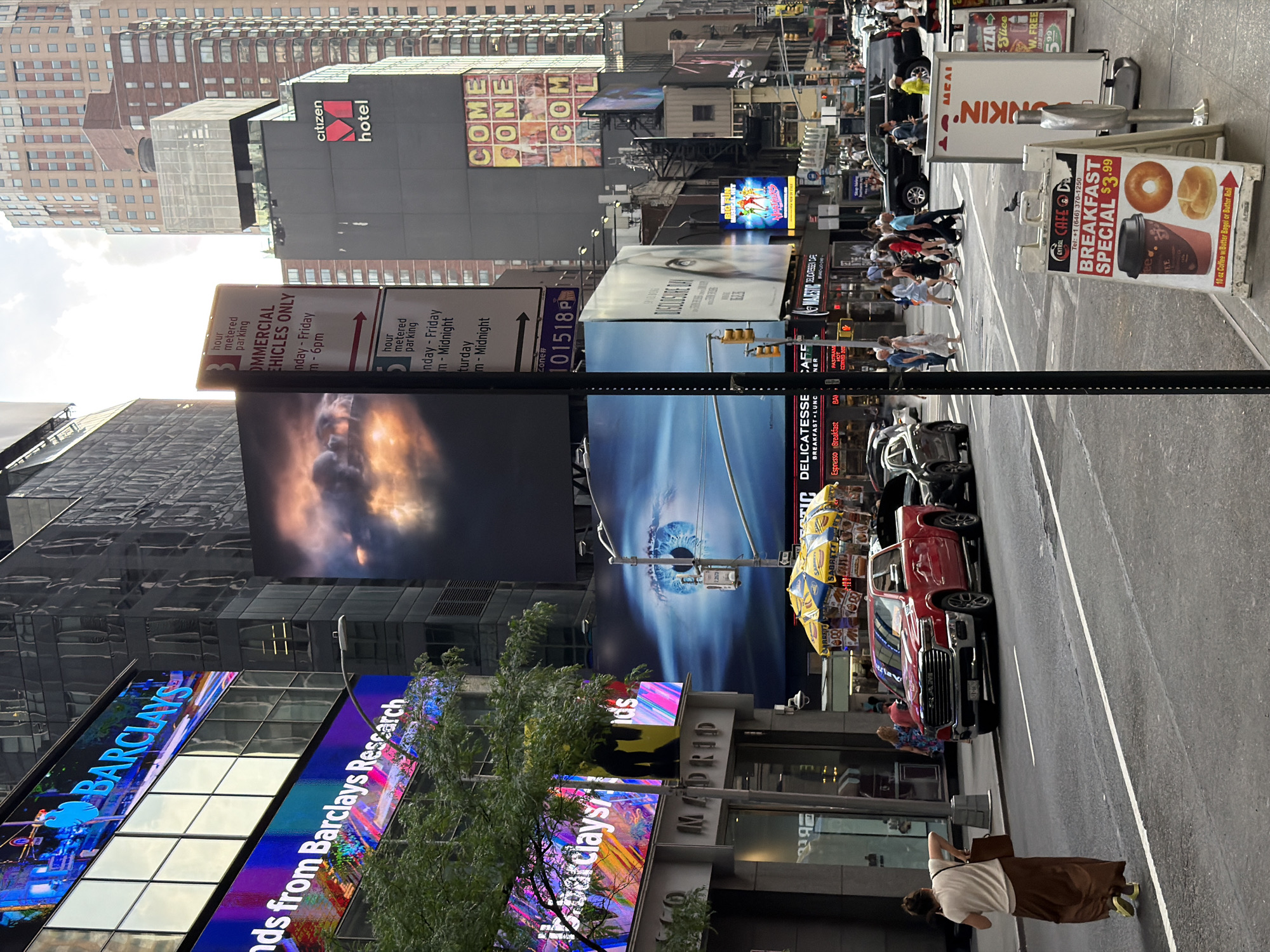 This image depicts a street scene in Times Square, New York City. The photograph shows a pedestrian crossing, with several people visible on the sidewalks and crosswalks. Signage is prominent, including electronic billboards for Barclays and Madrid, commercial vehicle parking signs, and a "Breakfast Special" sign advertising a café. Several vehicles, including cars and trucks, are on the street. The architecture is dense with tall buildings, including the Citizen Hotel. Bright advertising dominates the skyline. A woman with a brown skirt walks along the sidewalk toward the street.