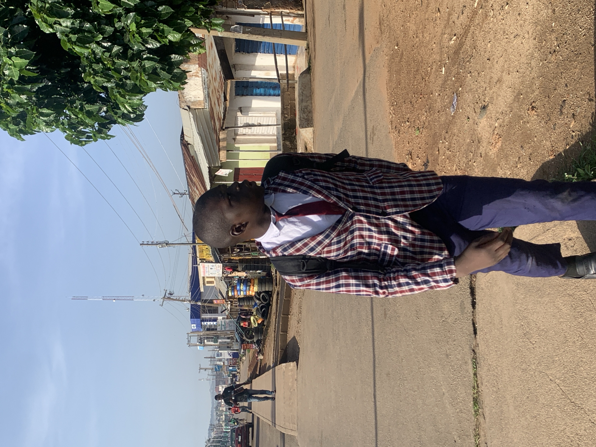 The image depicts a street scene in Jos, Nigeria, characterized by a paved road and a sidewalk. The foreground is dominated by a young male individual wearing a plaid jacket, a white shirt, a red tie, and blue trousers. He has a backpack on his shoulders and is looking to his right, away from the camera. His left hand is near his right knee.
The background reveals a busy street with several buildings lining one side. These buildings appear to be shops, some with corrugated iron roofs. Tires are stacked in front of one of the shops, and a tree with dense green foliage is visible on the right side of the frame. Another person is walking on the sidewalk in the mid-ground, heading away from the camera. Cars and a telecommunications tower are also present in the distance, suggesting an urban or semi-urban environment. The sky is clear and blue.