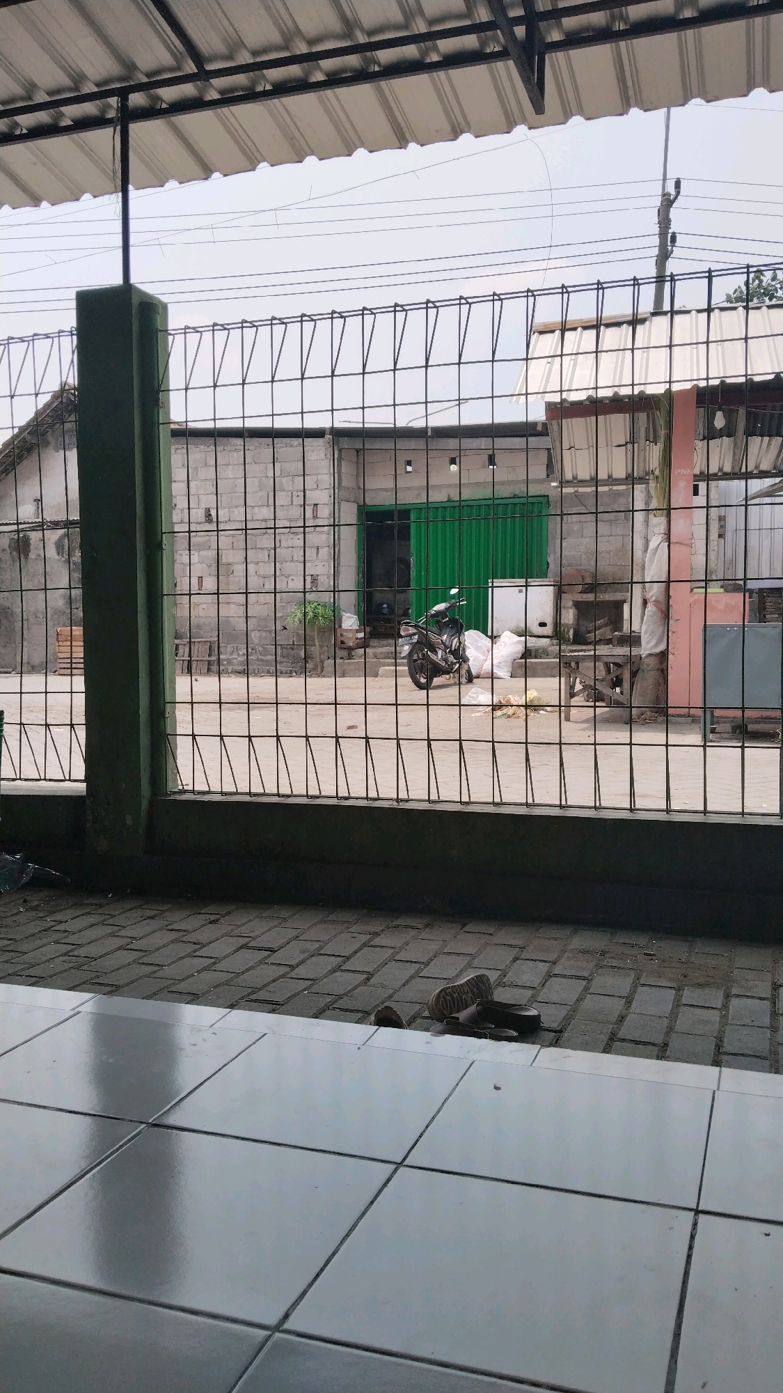 A low-angle medium shot captures a street scene in Losari, Indonesia, viewed through a metal fence. The foreground features a tiled floor with a slight sheen. The midground displays a paved area with several pairs of sandals scattered on the ground. Beyond the sandals, a black motorcycle is parked next to several large white sacks. Behind the motorcycle, a green metal shuttered door is visible, set into a wall constructed of grey cinder blocks. To the left of the motorcycle, a small green plant grows in front of a wooden structure. The background consists of a building with a corrugated metal roof and some overhead wires. The sky is overcast.