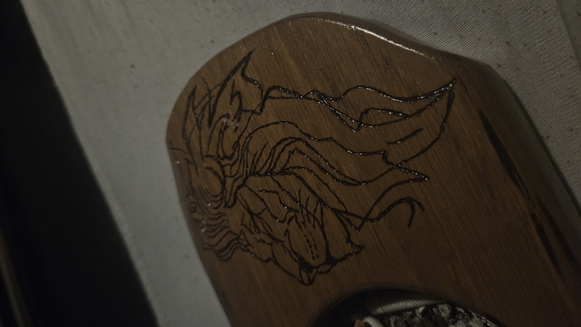 The image shows a close-up, angled shot of a piece of wood, possibly a carved plaque or a decorative item, with a design of a lion's head etched into its surface. The etching is done in black, creating a stark contrast against the brown wood. The wood appears to have a glossy finish, reflecting light in small, scattered patches, some of which appear to be water droplets. The lion's mane is depicted with flowing lines radiating from its head. The object is placed on a textured, light-colored surface, possibly a fabric tablecloth or a similar material. The background is dark and out of focus, with a blurred horizontal line suggesting a separation between surfaces or lighting changes. The setting is indeterminate and no specific urban location can be identified.