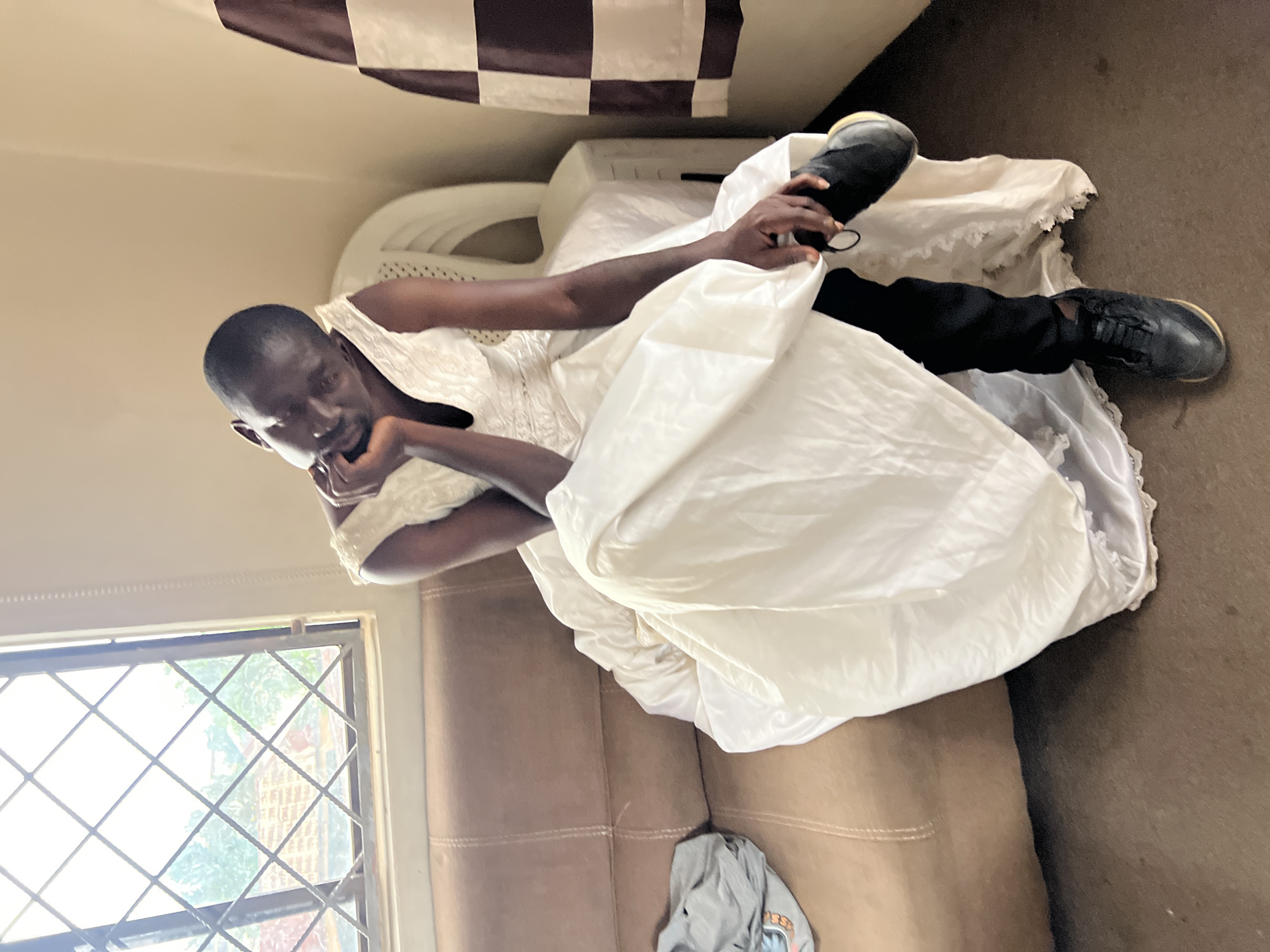 A person, appearing to be male based on facial features and visible arms, is seated on a brown leather couch, wearing a white wedding dress and black pants and shoes. The person is leaning on their hand, with their head tilted to the side, and their right foot is raised, with a black shoe held in their hand. A grey t-shirt is draped on the couch to the left of the person. The background includes a white plastic chair, a cream-colored wall, and a window with a diamond-patterned metal grate, through which trees and rooftops are visible. A checkered curtain hangs on the right wall. The floor is a dark brown, textured surface. The scene appears to be an indoor setting, likely a room within a residential building in Jos, Nigeria.