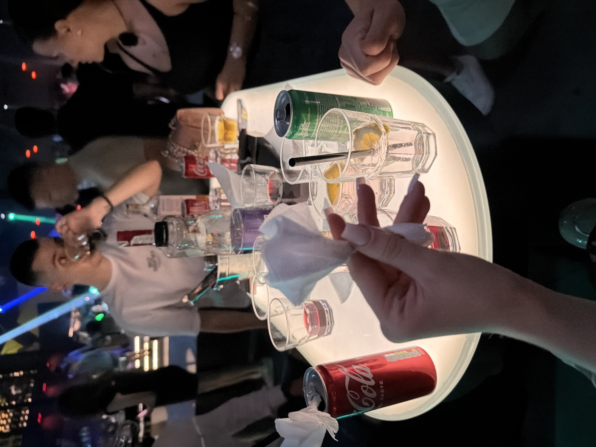 The image depicts a dimly lit indoor environment, likely a nightclub or bar, with a table in the foreground illuminated by an internal light source. On the table, there are several drinks, including a red Coca-Cola can, a green Sprite can, and various glasses containing liquids and ice. One glass has a straw and lemon slices. A hand in the foreground is holding a white tissue. In the background, several individuals are visible, some blurred due to movement or lighting conditions. The overall atmosphere suggests a social gathering or nightlife scene. The location is identified as Varna, Bulgaria.