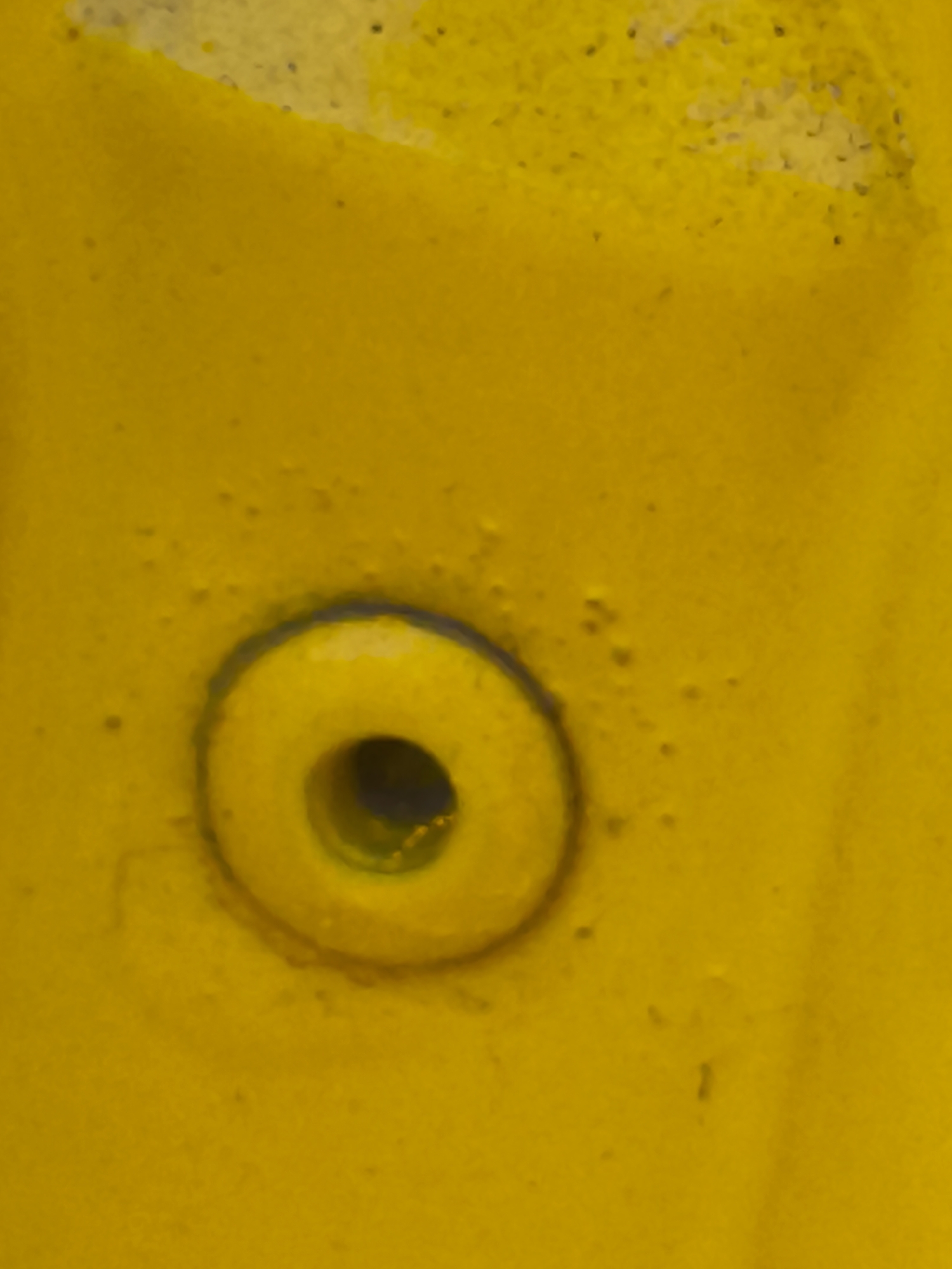 This image is a close-up shot of a yellow, textured surface with a circular indentation. The indentation has a raised rim and a dark opening in the center, suggesting it might be a bolt head, a recessed button, or a similar fixture. The surface appears to be outdoors, given the slight dappling of light and shadow, and the texture suggests it could be a metal or plastic material, possibly painted. There are small dark specks scattered across the yellow surface, which could be dirt, debris, or part of the material's texture. The lighting suggests it is daytime, but the weather is not discernible. There is no text visible in the image. The context provided places this scene in Nowy Dwór Mazowiecki, Poland.