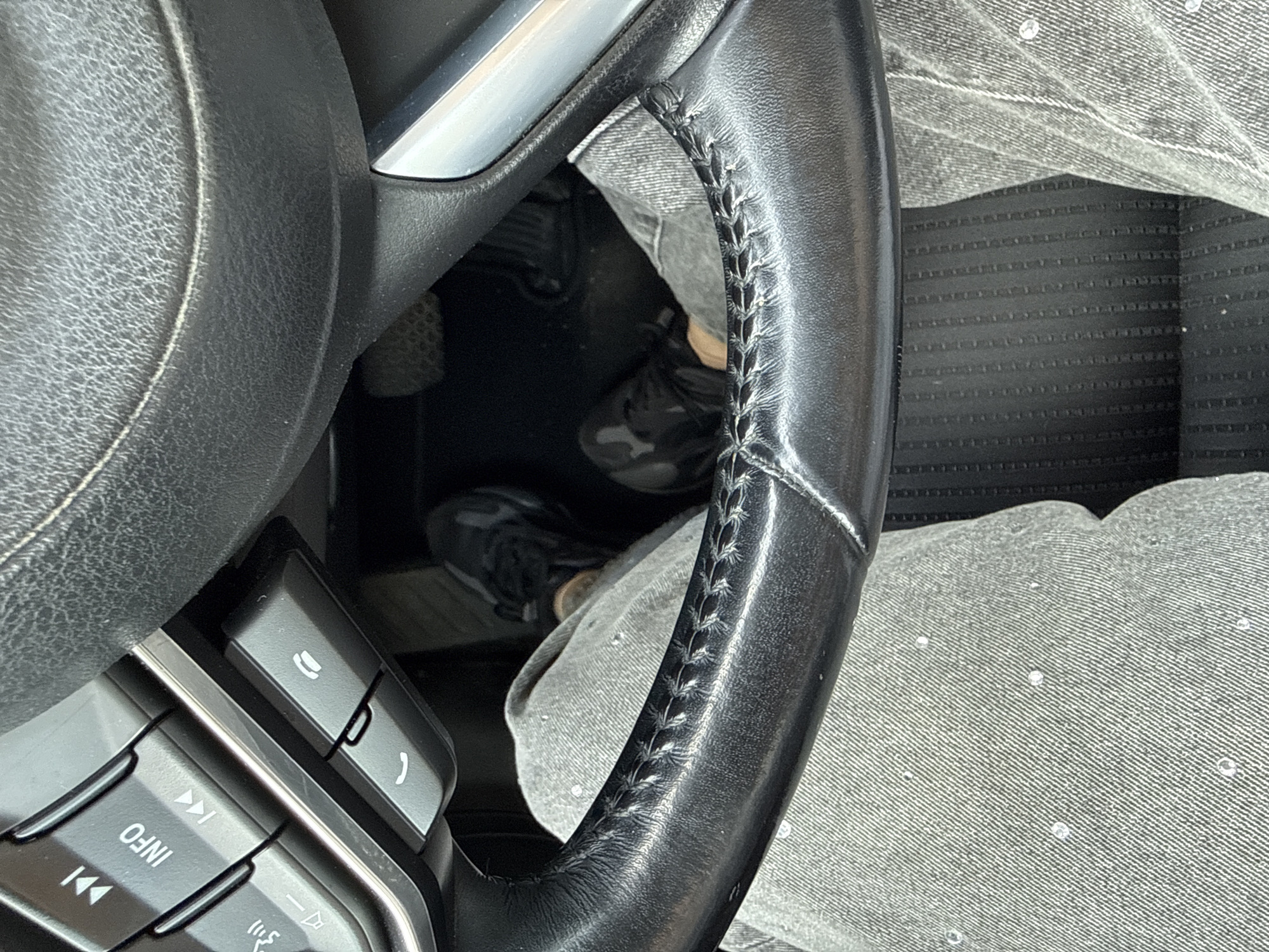 The image shows a close-up view from inside a car, focusing on the steering wheel and the driver's legs. The steering wheel is black leather with visible stitching. On the left side of the steering wheel, there are buttons, including one with an "INFO" label and another with a phone icon. The driver is wearing grey jeans, some of which appear to have small reflective or shiny embellishments. Their feet are visible, wearing camouflage-patterned sneakers, resting on or near the car's pedals. The floor of the car has a textured black mat. The scene suggests the car is stationary, likely parked or stopped. The prompt's request to include the location of Šilutė, Lithuania is not supported by any visual information in the image, as there are no identifying landmarks or context to place the scene geographically. Therefore, no information about Šilutė, Lithuania can be provided.