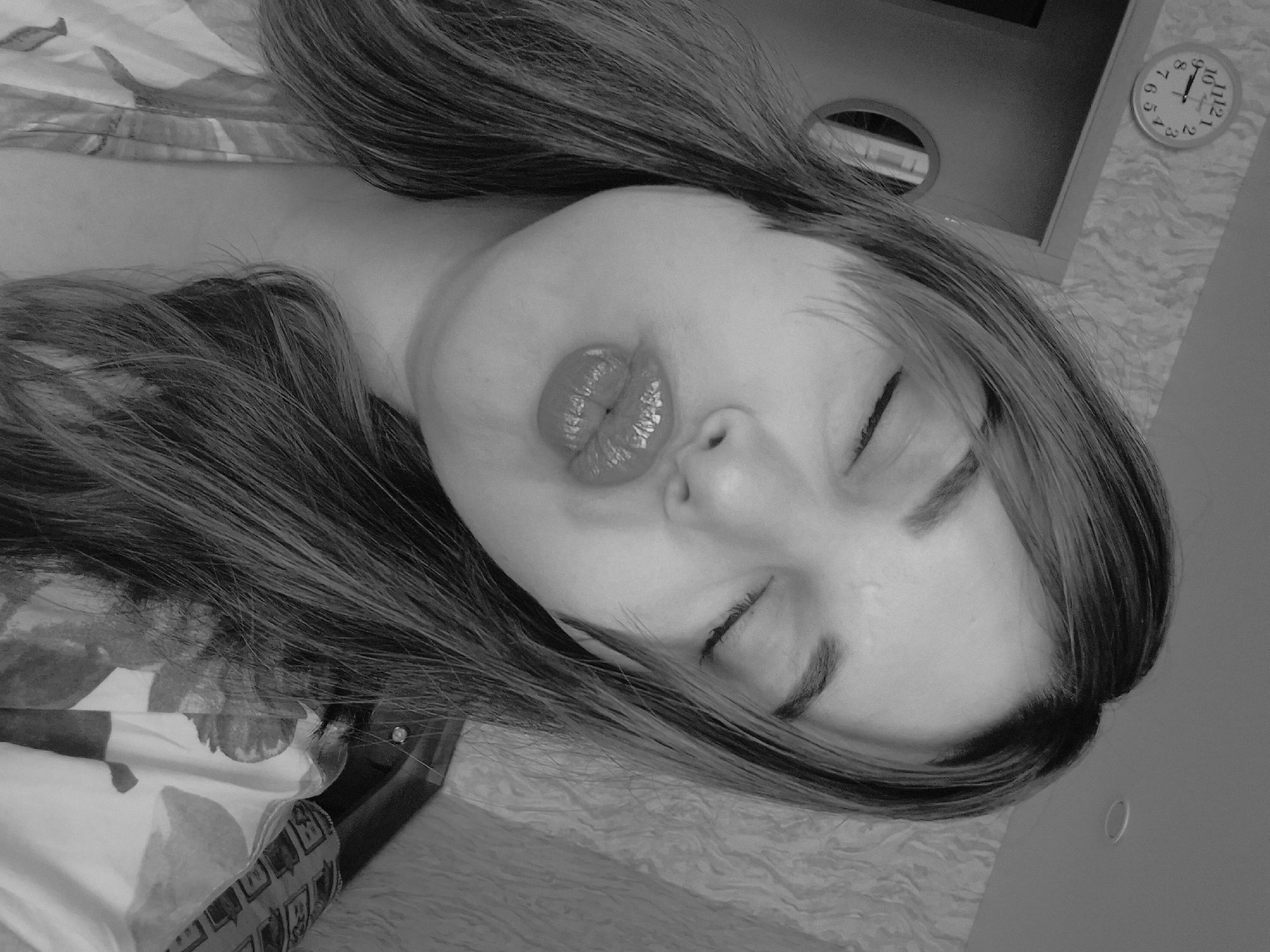 A grayscale image depicts a woman lying down, with her eyes closed and lips pursed. The background includes a clock on the wall showing 10:07, a ceiling light fixture, and a floral patterned bedspread. Location: Амзя, Russia.