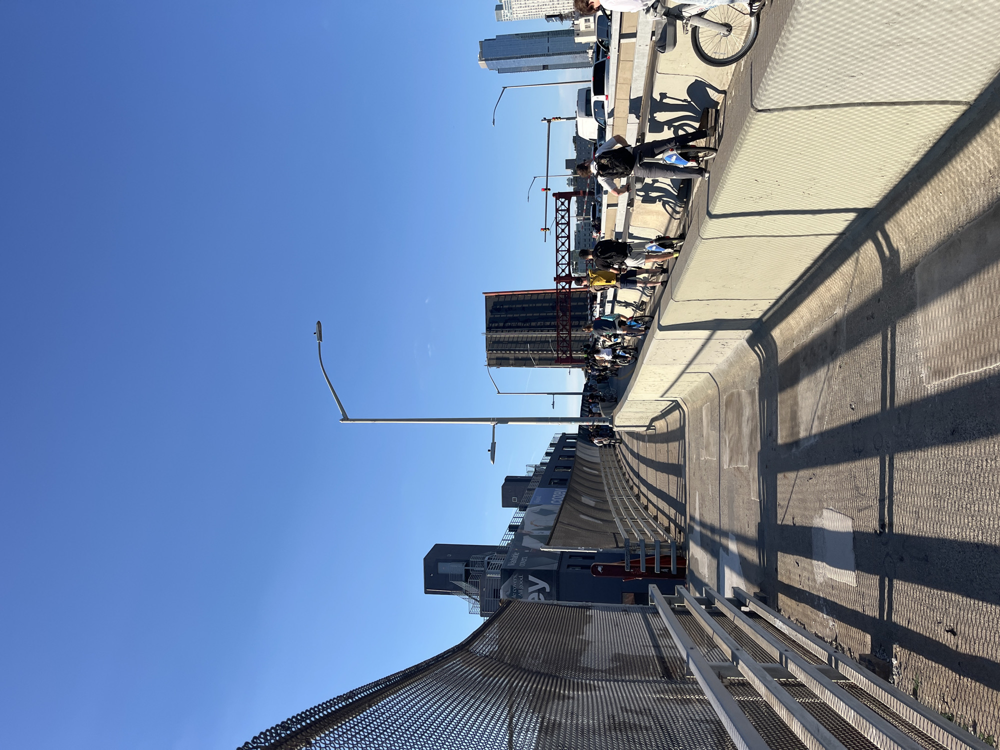 A bright, clear blue sky stretches above a pedestrian and bicycle path on a bridge in New York City. The path is lined with a metal fence on the left, casting long, striped shadows on the concrete walkway. On the right, a concrete barrier separates the path from a roadway below, where cars and vans are visible. A group of people are riding bicycles and electric scooters on the path, some with backpacks. In the background, a tall, modern building with a red accent is visible, along with other high-rise structures and a drawbridge. The scene depicts a sunny day on a bustling urban bridge.