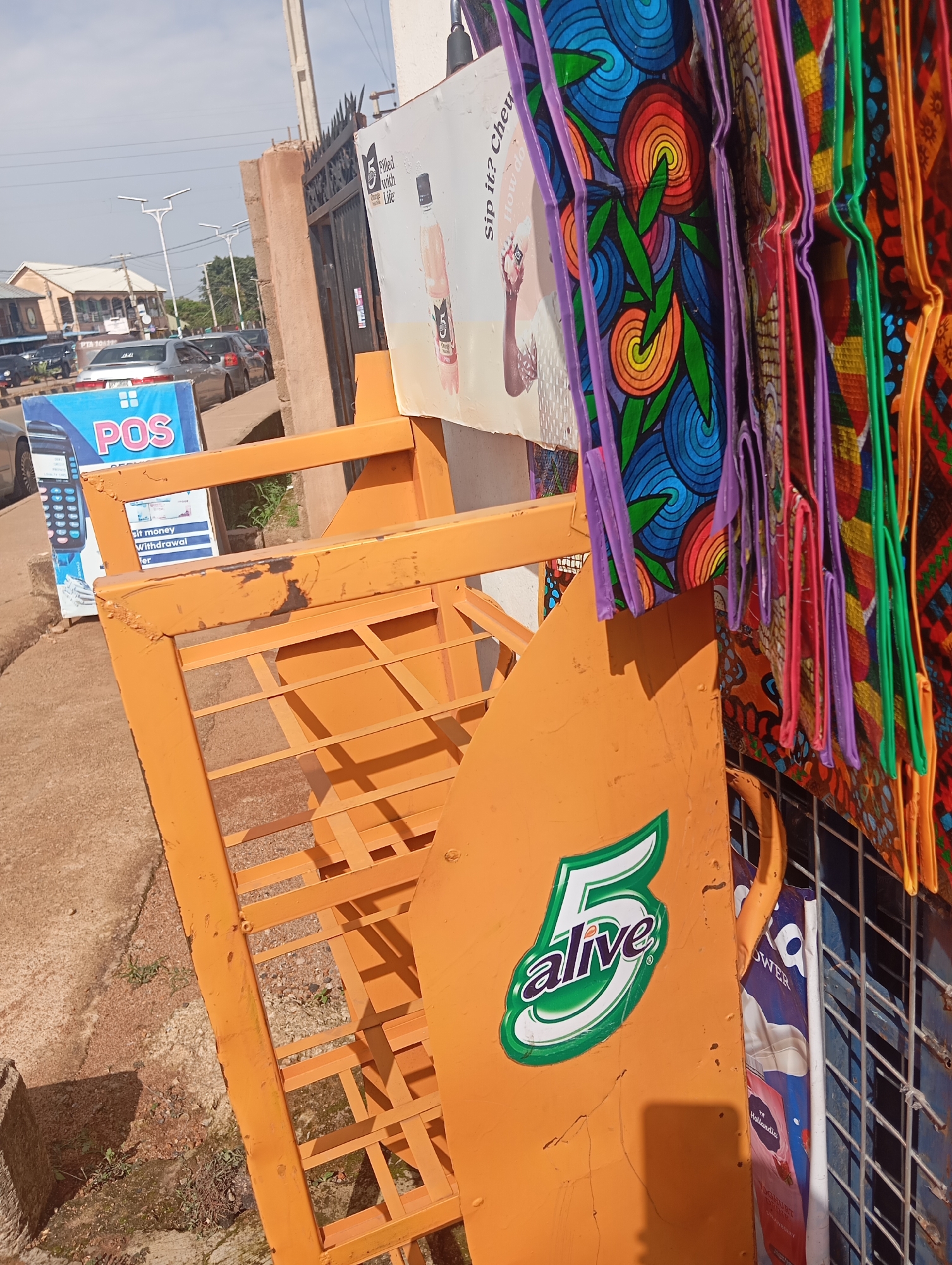 The image shows an outdoor scene in Gura Top, Nigeria, likely during the day under a clear sky. In the foreground, an orange metal rack stands diagonally, suggesting it is part of a stall or display. Attached to the rack is a large orange sign featuring a green and white "5 alive" logo. To the left of the rack, a blue and white sign advertises "POS" services, including "sit money" and "Withdrawal." Behind this, a row of colorful, patterned bags, possibly for sale, hangs from a metal grid. The bags display vibrant geometric and floral designs in shades of blue, red, orange, green, purple, and yellow.
Further back, a fence with decorative metalwork separates the stall area from a street with light traffic. Cars are visible on the road, and beyond them, buildings with distinct architectural styles, including what appears to be a mosque with a minaret, are present. Streetlights are also visible, indicating a developed urban or semi-urban environment. The overall impression is a bustling street-side market or commercial area in Nigeria. No people are clearly visible in the frame.