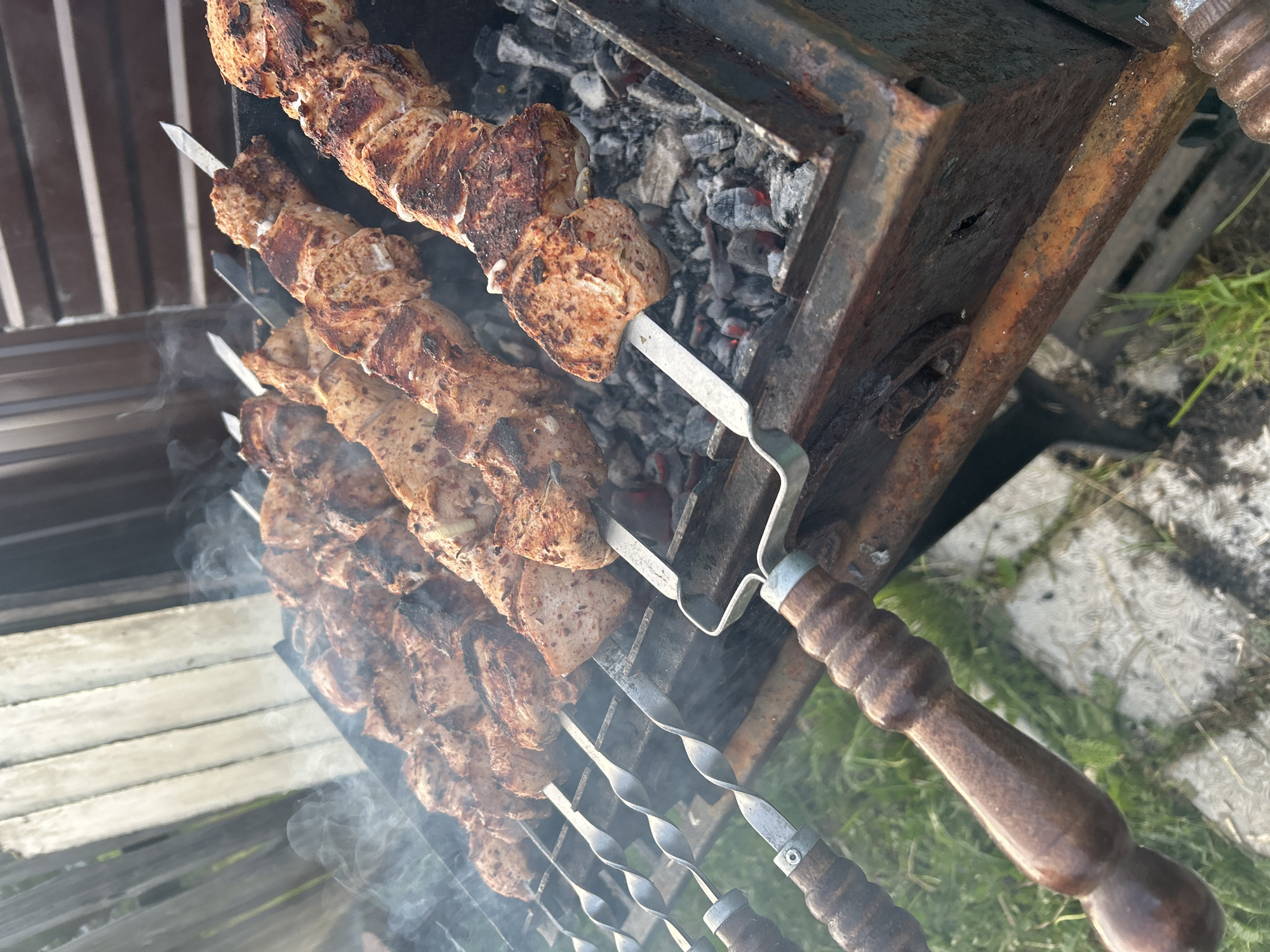 The image shows shish kebabs being grilled on a brazier. The skewers are metal and twisted, with wooden handles. The brazier is rusty and contains hot coals. The scene appears to be outdoors, with grass and concrete visible in the background. The location is inferred to be in Ольховка, Russia based on the prompt.