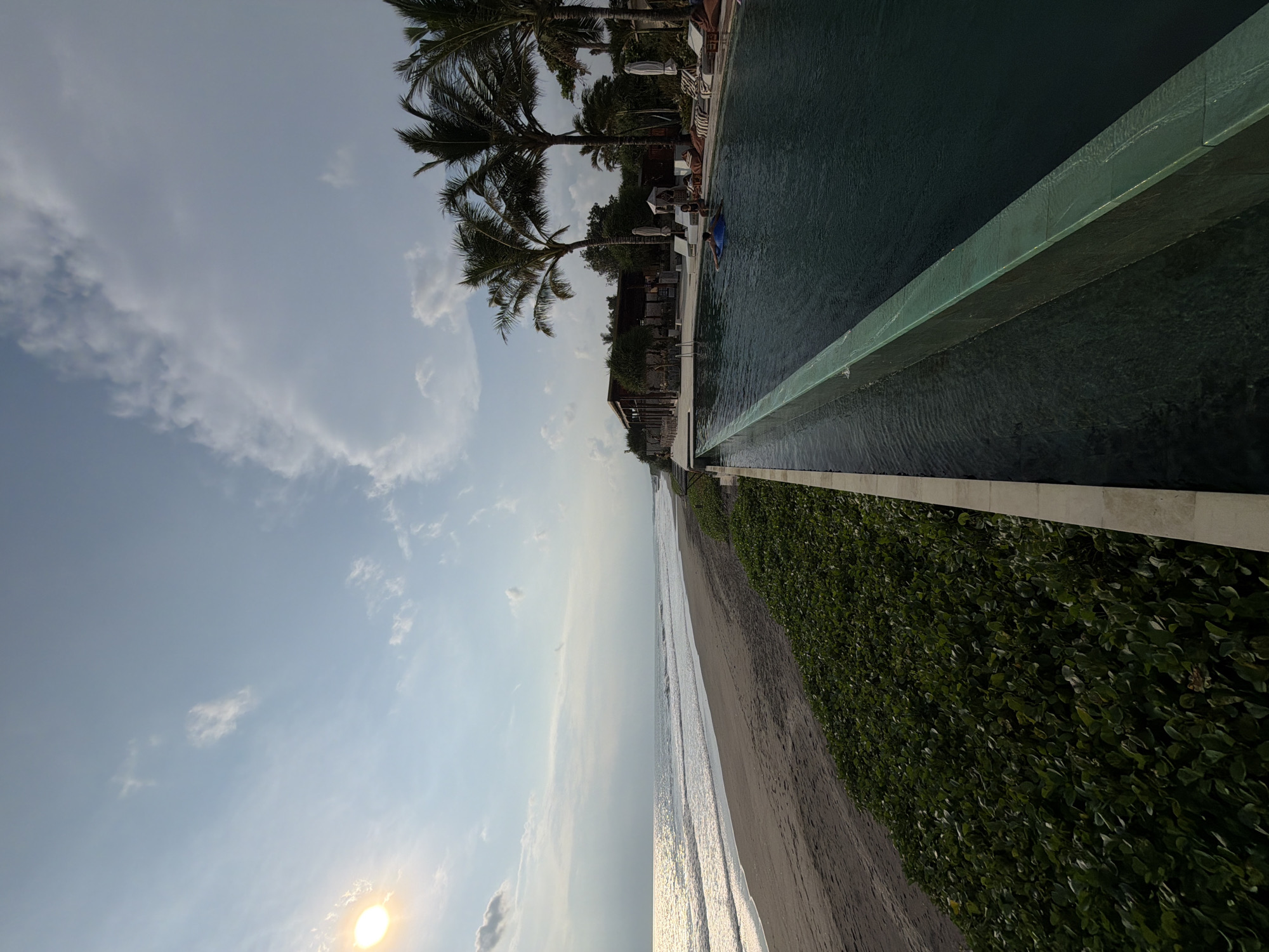 The image displays a coastal scene in Belalang, Indonesia, captured during late afternoon or early morning. A wide expanse of dark grey sand beach extends from the left midground towards the right, meeting the dark greyish-blue ocean where white waves break repeatedly. The sun, a bright yellow sphere, is low on the horizon in the upper left, casting a strong glare across the ocean's surface and illuminating the underside of scattered white clouds in the light blue sky.
Running parallel to the beach, a dense strip of low-lying green groundcover separates the sand from a resort or hotel complex. The foreground is dominated by a dark-water infinity-edge swimming pool, its greenish-blue textured wall forming a waterfall effect into a lower trough. One individual, partially submerged, floats in the pool on a blue inflatable ring, facing towards the right.
Beyond the pool, a paved area features multiple white lounge chairs, some with striped cushions, and several large, dark beanbag-style chairs. Two individuals are seated on these chairs near the pool's edge, partially obscured by open white beach umbrellas and tropical foliage. Further back, numerous tall palm trees line the property, and several resort buildings are visible, including a prominent open-air structure with a dark roof and wooden pillars. A small, white conical cabana or tent is also present in the lounge area.